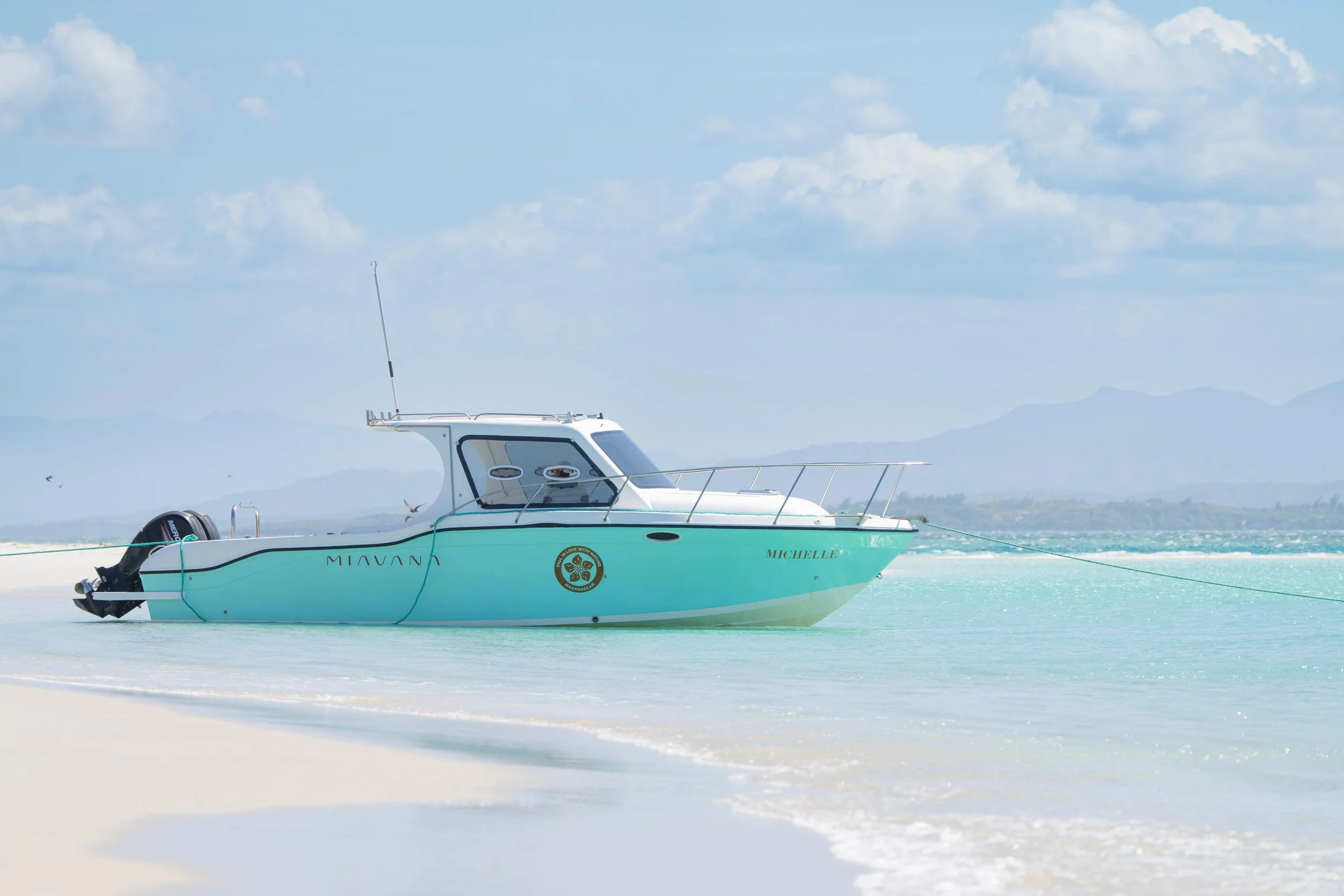 A turquoise motorboat named MICHELLE anchored on calm, shallow waters near a white sandy beach with distant mountains and a partly cloudy sky.