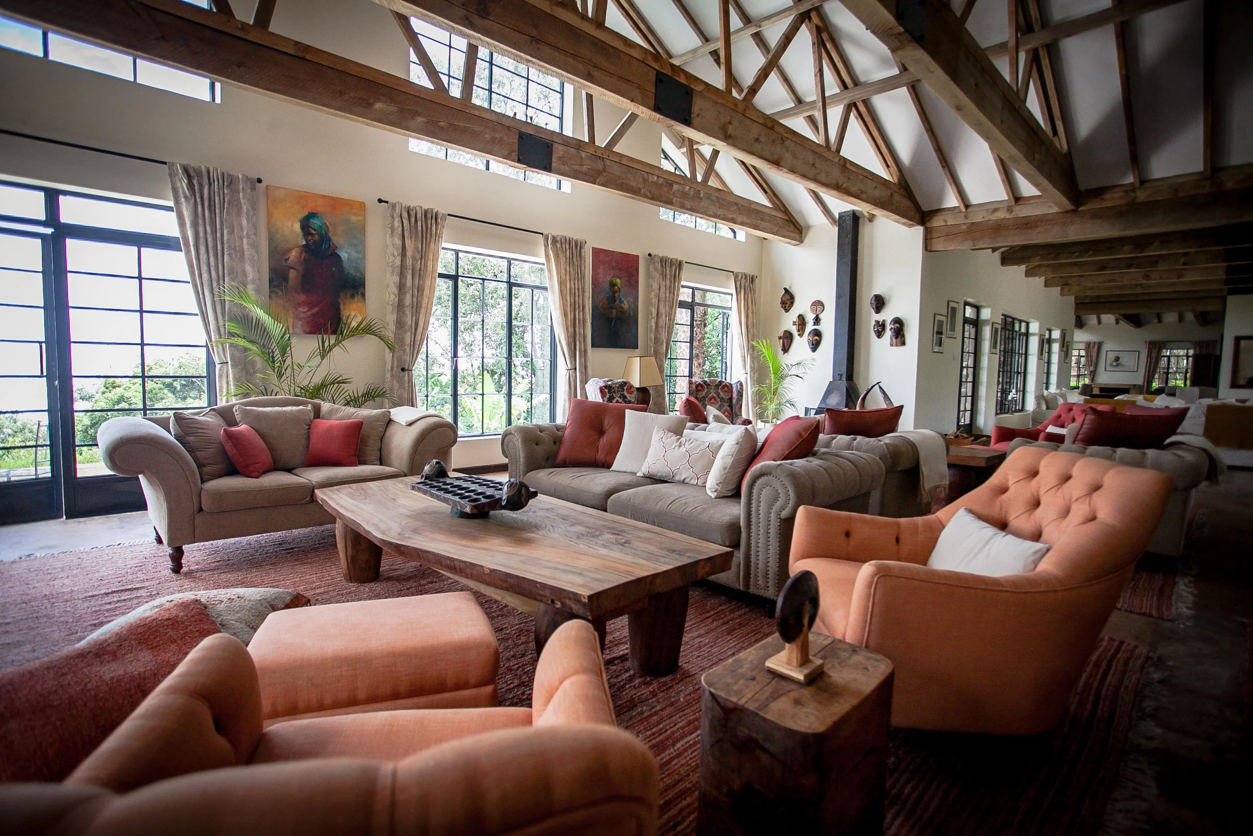Spacious living room with beige sofas and peach armchairs, wooden coffee table, large windows with patterned curtains, colorful artwork and tribal masks on the walls, potted plants, exposed wooden beams, and a mix of modern and rustic decor.