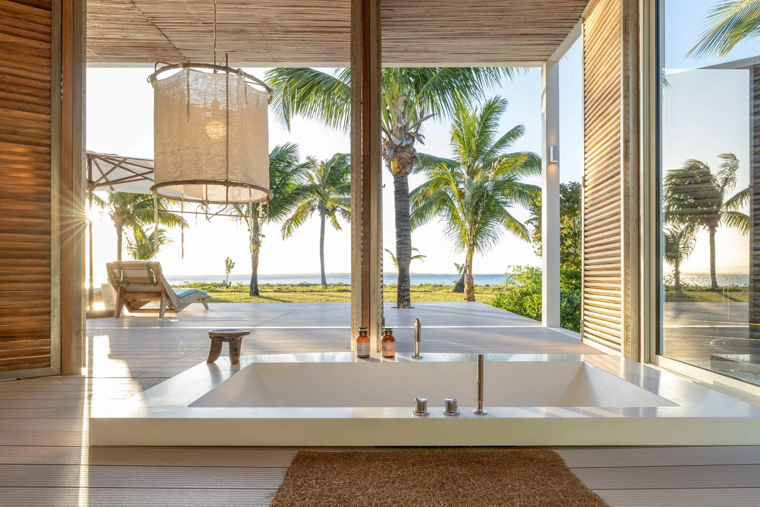 Indoor bathroom with a built-in bathtub looking out to a tropical outdoor patio with palm trees and a view of the beach