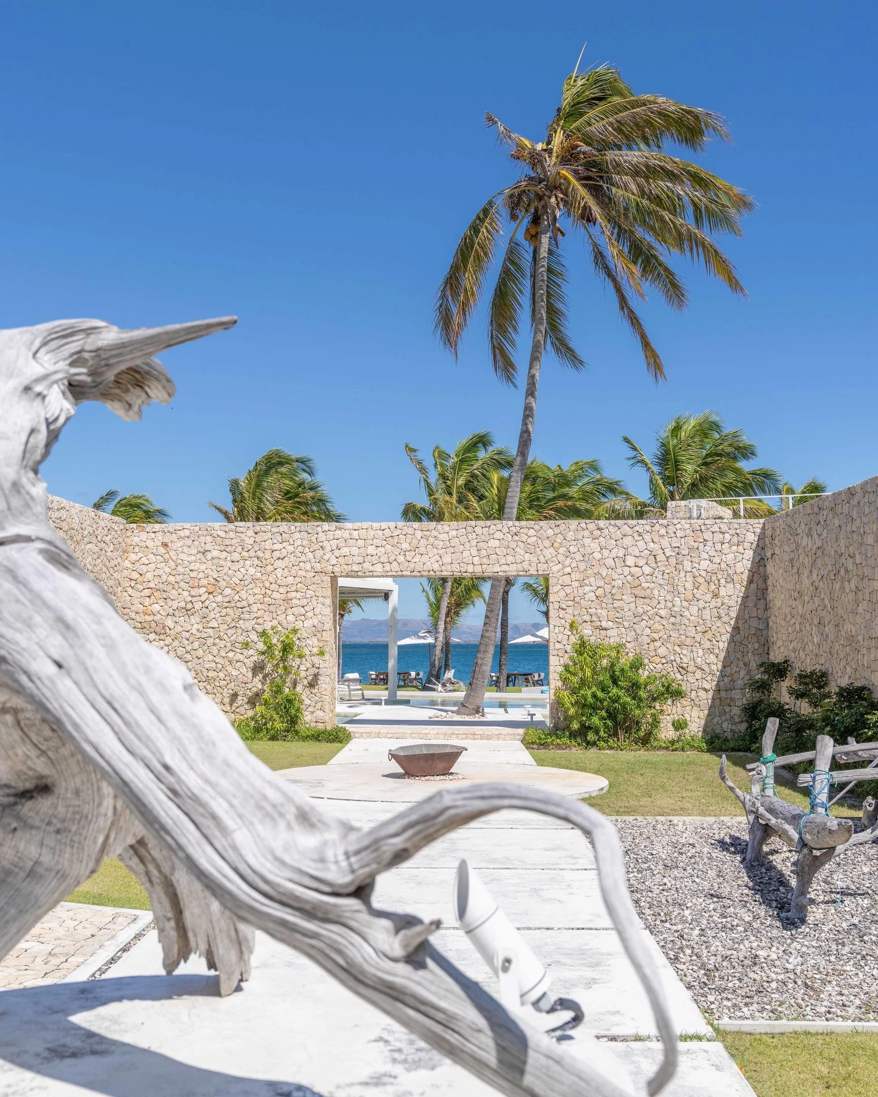 Tropical beach scene with palm trees, stone wall, ocean view, and blue sky, with decorative ocean-themed sculpture in the foreground.