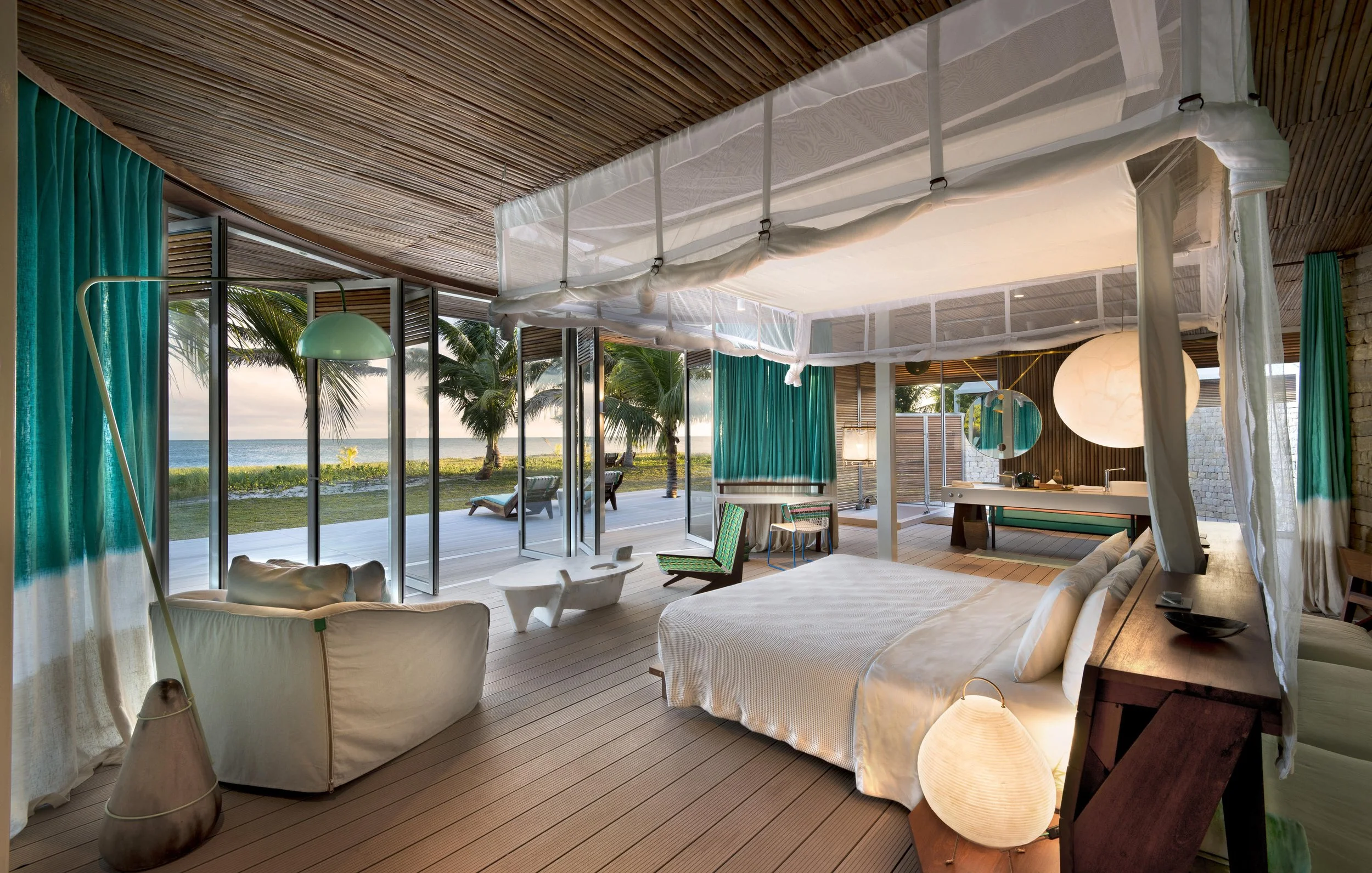 Luxurious beachfront hotel room with a sea view, white bed, seating area, outdoor deck with lounge chairs, palm trees, and ocean in the background.