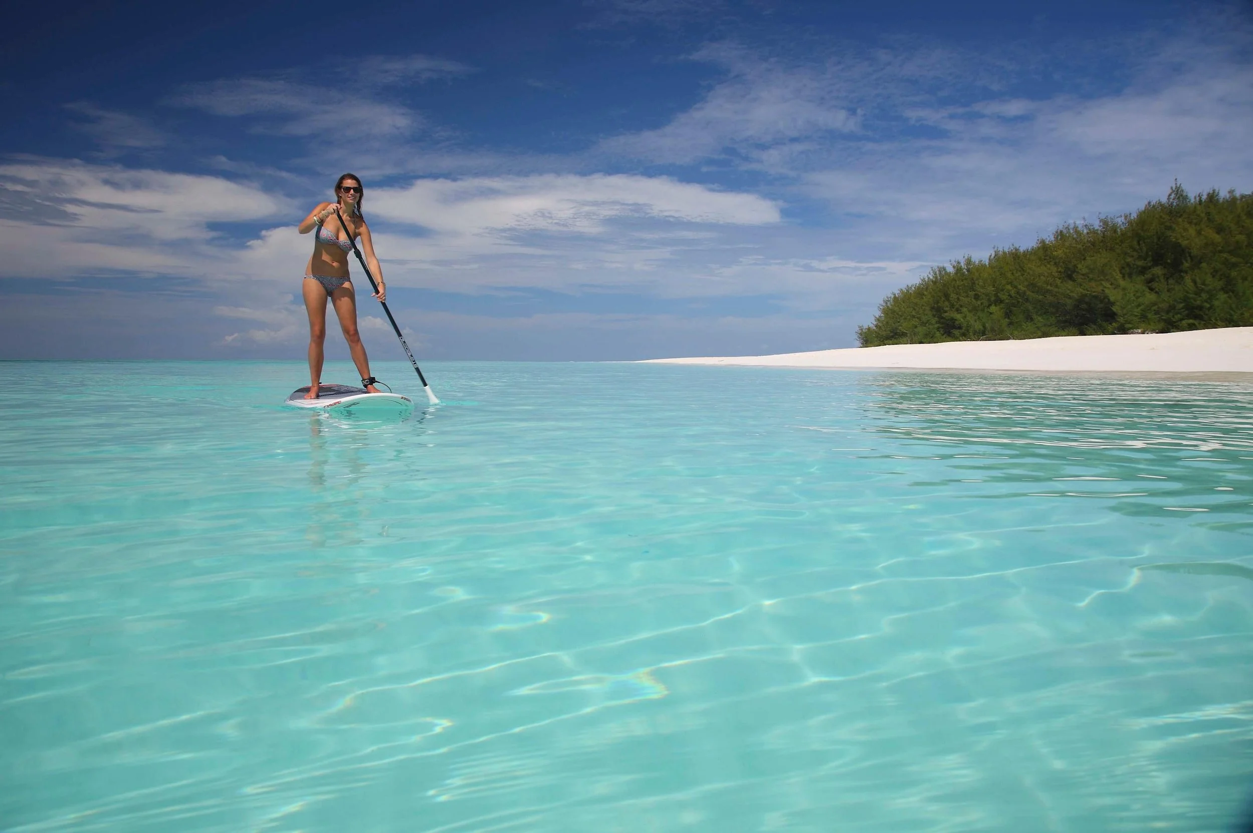 Woman paddleboarding in clear turquoise water near a sandy beach with trees and blue sky