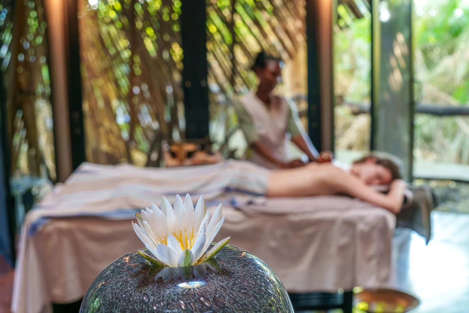 Close-up of a white water lily floating on a dark stone bowl, with a massage being performed on a woman lying face down on a massage table in the background.