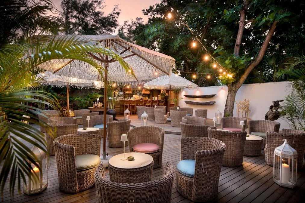 An outdoor patio area with wicker chairs, small tables, and umbrellas, decorated with string lights, lanterns, and lush green plants at sunset.