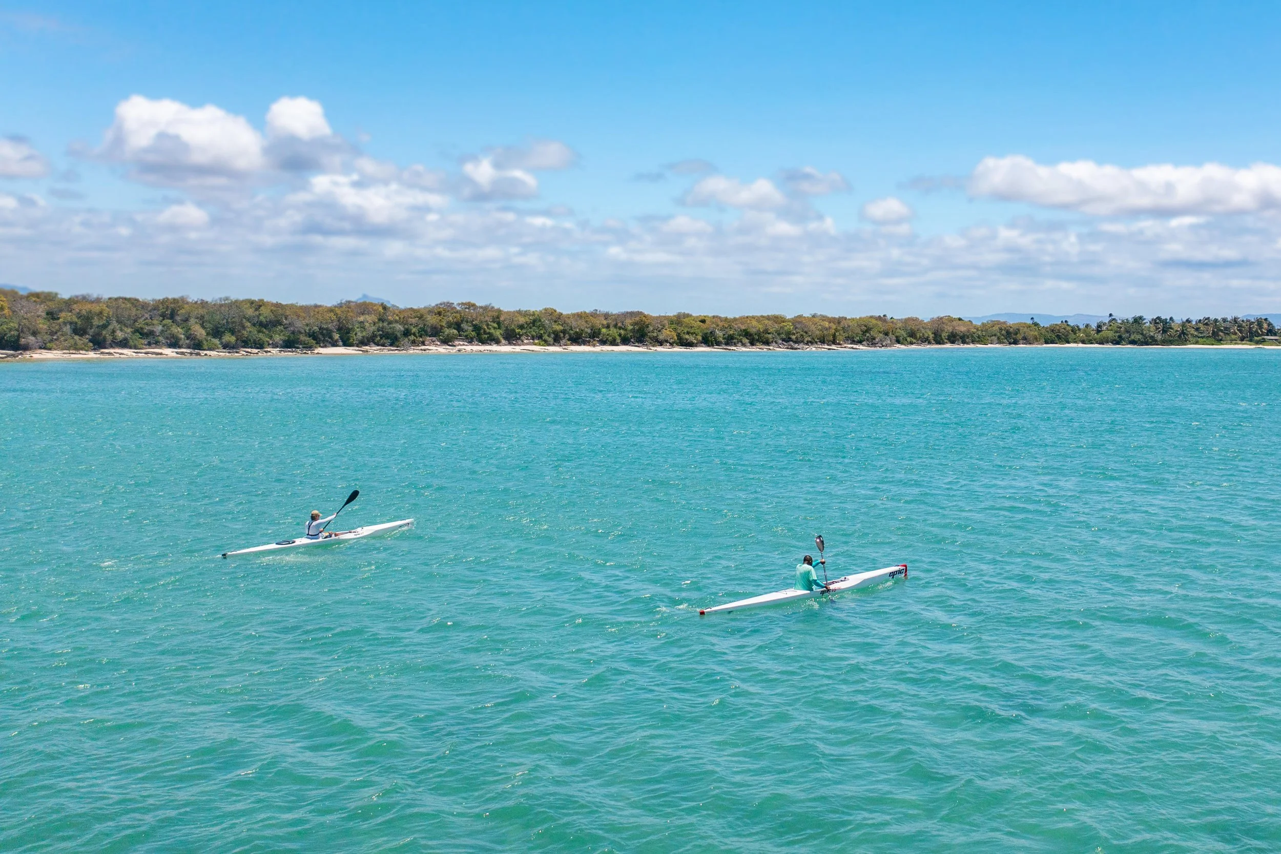 Two people kayaking on calm turquoise water with a sandy shoreline and trees in the background under a partly cloudy sky.