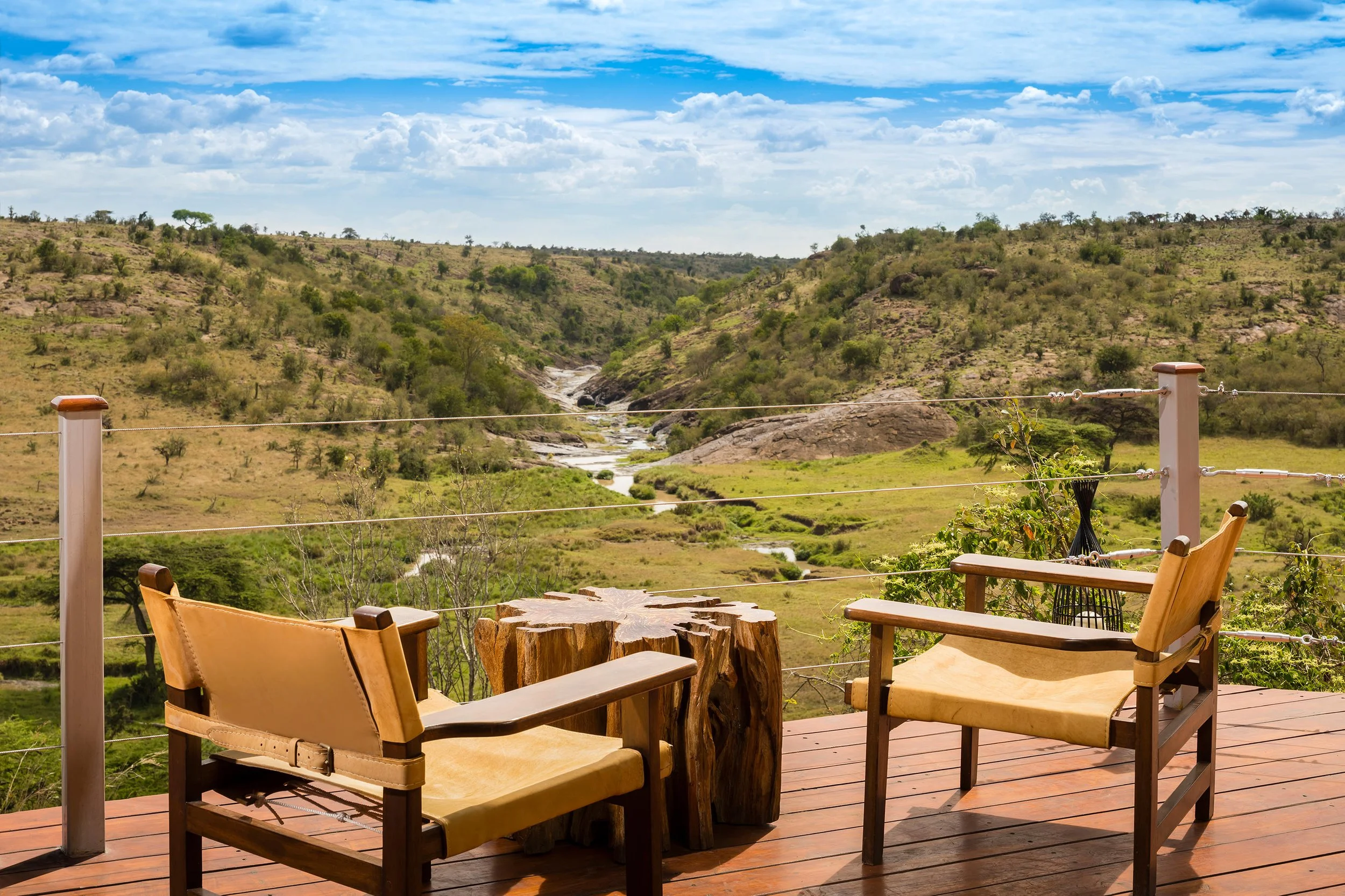 A wooden deck with two chairs facing a scenic view of rolling hills, a river, and scattered trees under a blue sky with clouds.