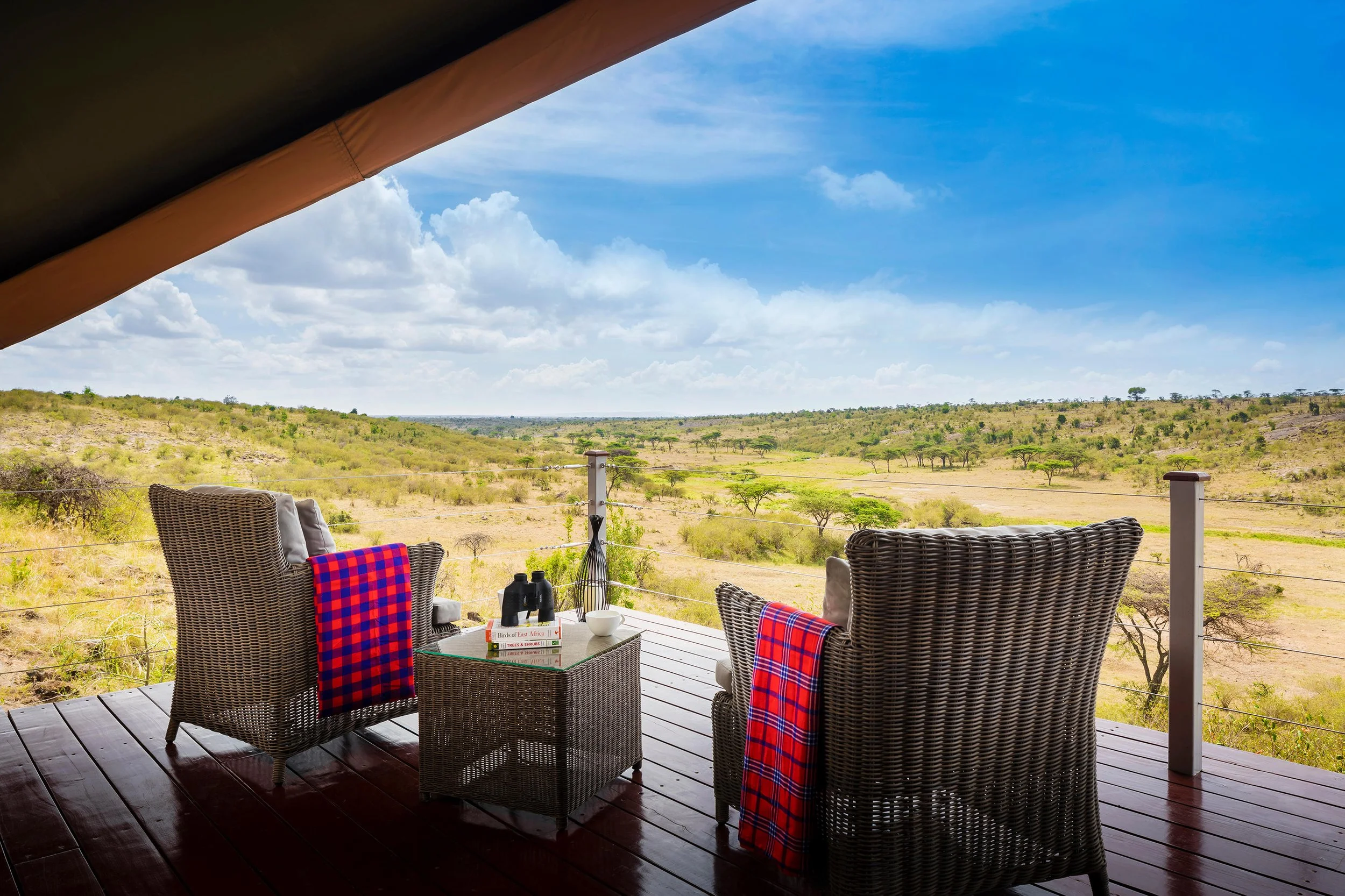 Outdoor patio with wicker chairs, a small table with binoculars, a bowl, and books, overlooking a savannah landscape with scattered trees under a partly cloudy sky.