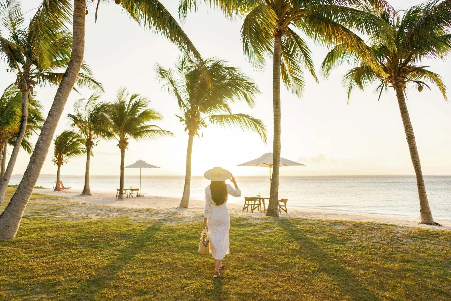 A woman walking on the grass near the beach during sunset, wearing a white dress and a wide-brimmed hat, carrying a bag, with palm trees and beach umbrellas in the background.