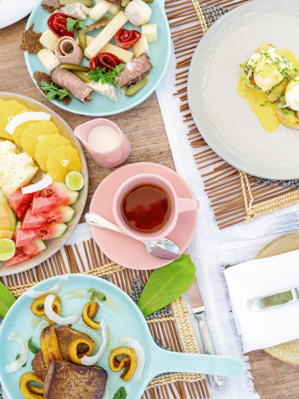 A table set with various foods including a fruit platter with watermelon, oranges, and green fruit slices, a plate of fried chicken with yellow peppers and onions, a cheese and vegetable plate, and a plate of cold cuts and cheese with pickles and tom