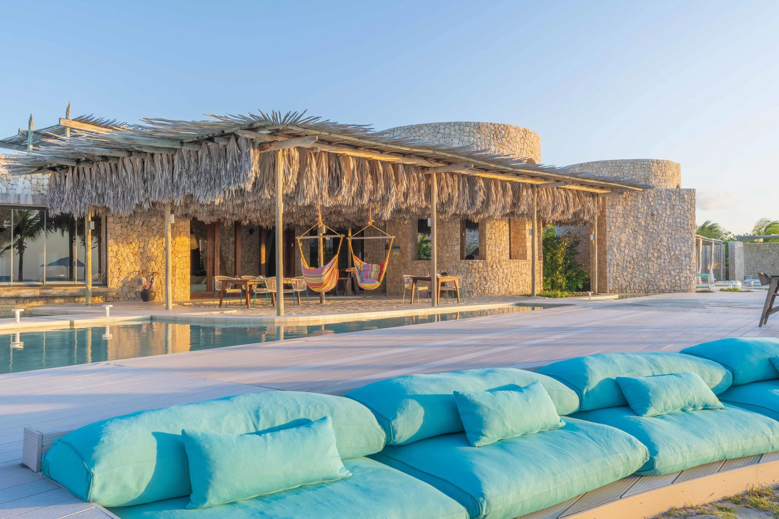 Luxury villa with thatched roof, hanging chairs, pool, inflatable lounge chairs, and clear sky.