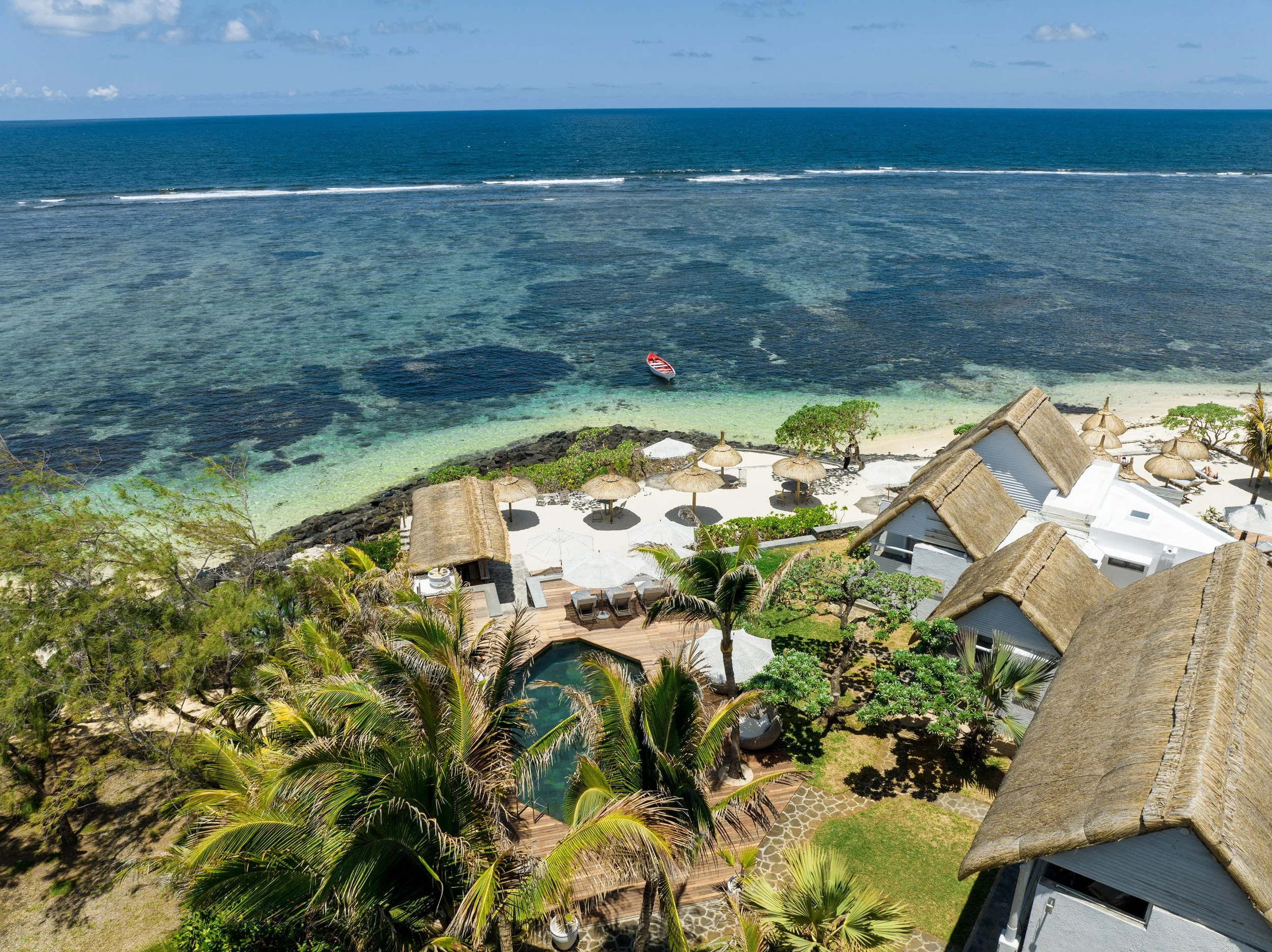 Aerial view of a beach resort with thatched-roof villas, palm trees, a swimming pool, and a sandy beach with umbrellas and lounge chairs, overlooking the ocean with a boat nearby.