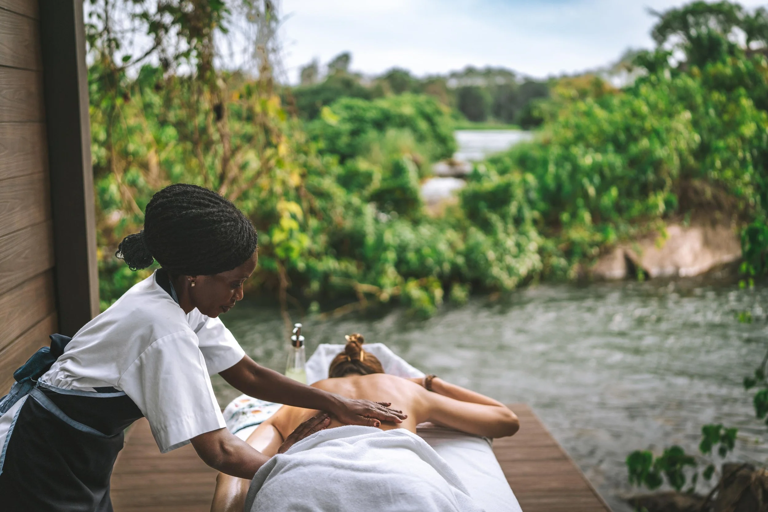A woman receives a massage outdoors on a wooden deck beside a river, surrounded by lush green trees.