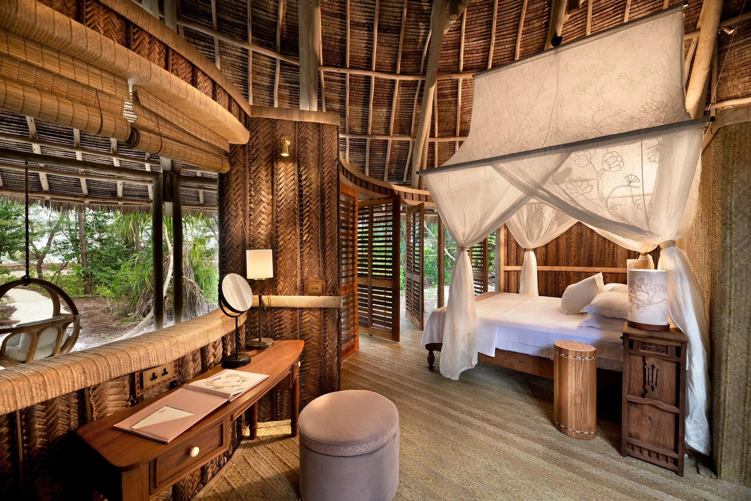 A rustic bedroom with wooden walls and a four-poster bed with a white canopy, surrounded by tropical greenery visible through open wooden shutters.