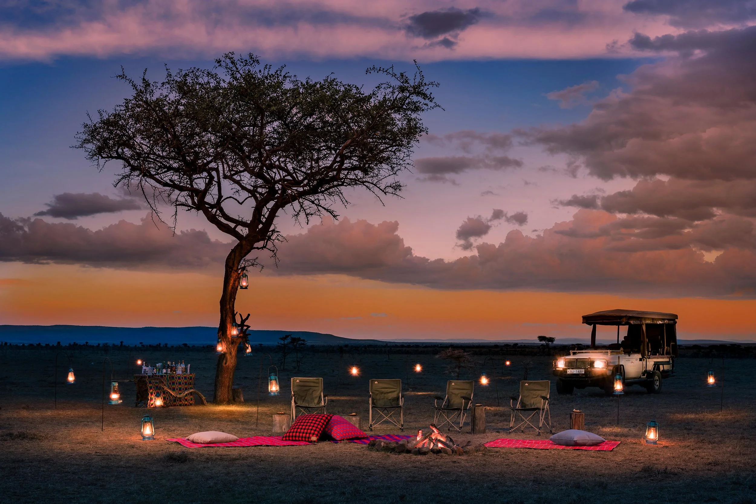 Sunset scene with a large tree decorated with hanging lanterns, surrounded by lanterns on the ground, with a picnic setup including chairs, a fire pit, pillows, and a lantern-lit table, and a vehicle parked nearby in an open field.