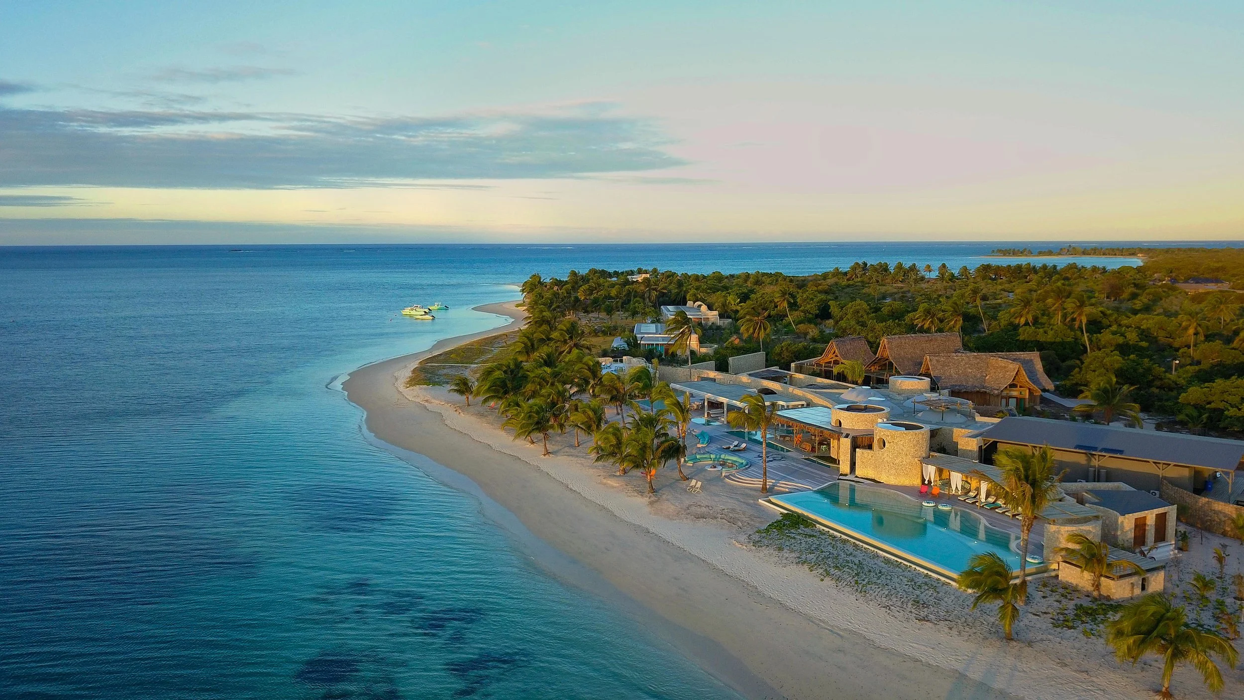 Aerial view of a tropical beach resort with a large swimming pool, sandy shoreline, and lush greenery with accommodations in the background, near the ocean.