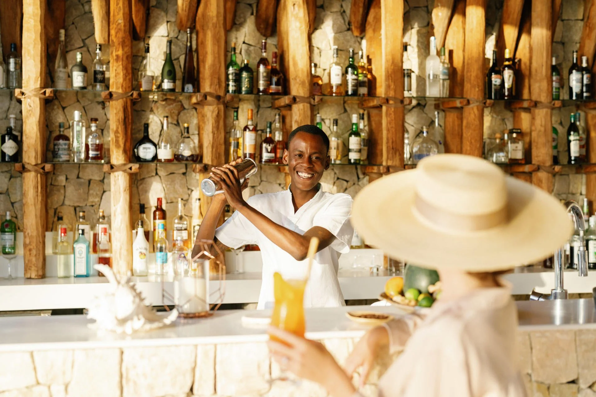 A smiling bartender shaking a cocktail shaker behind a bar with bottles of liquor on shelves, while a woman in a sunhat holds a drink with a straw.