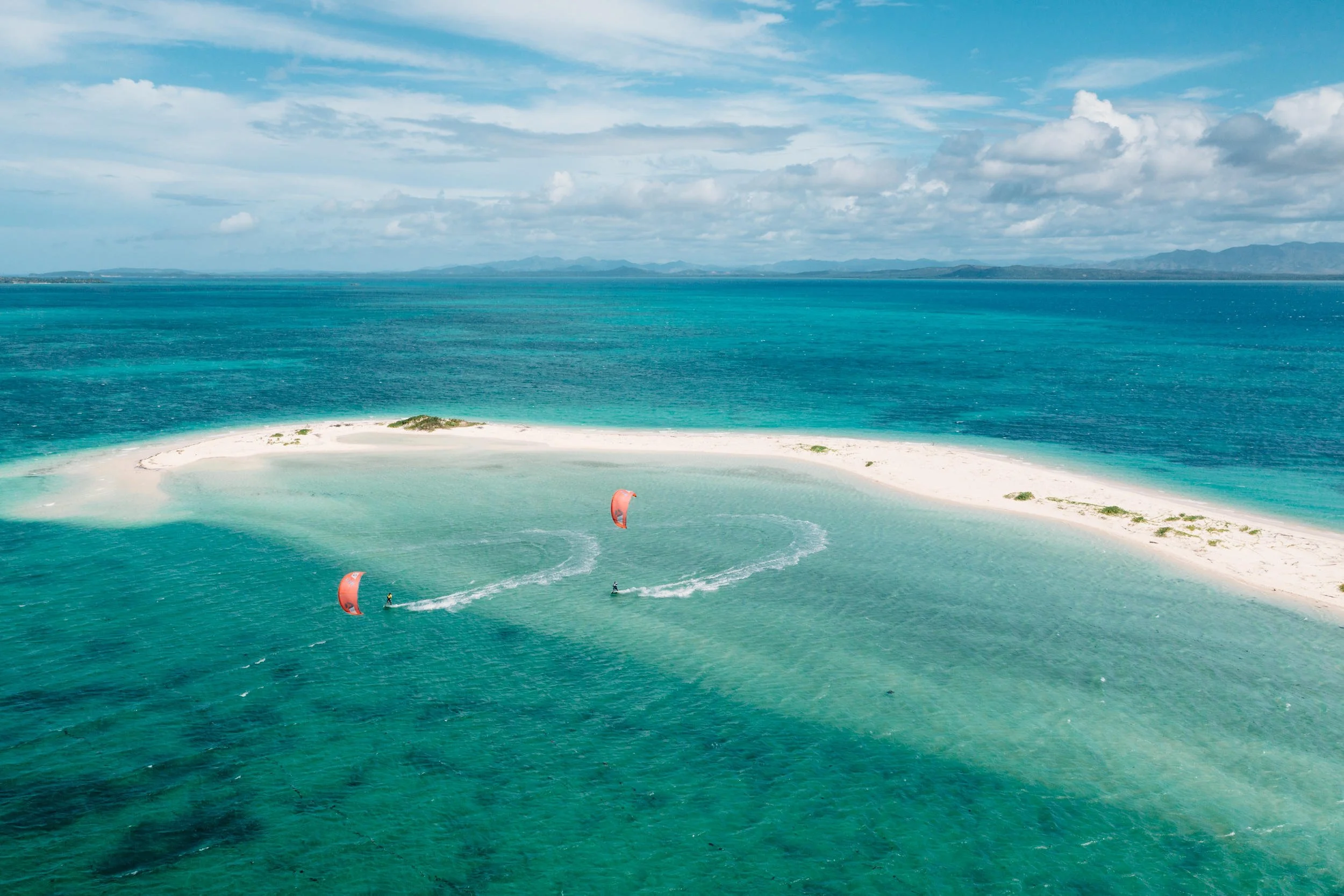 Two people kite surfing near a sandbar in clear blue ocean waters under partly cloudy sky.