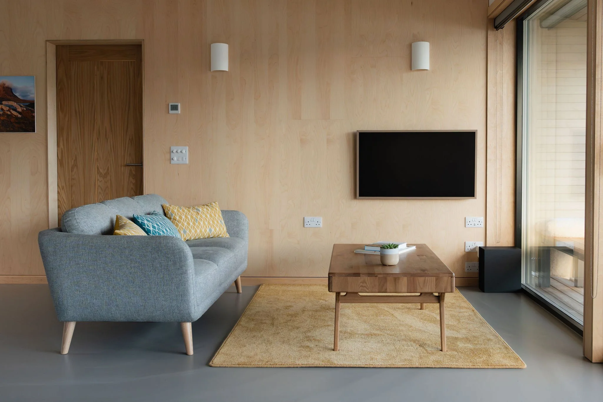 Modern living room with light wood paneled walls, a gray upholstered sofa with yellow and blue patterned pillows, a wooden coffee table with a potted plant and books, a flat-screen TV mounted on the wall, large windows with sliding glass doors, and a yellow area rug on the floor.