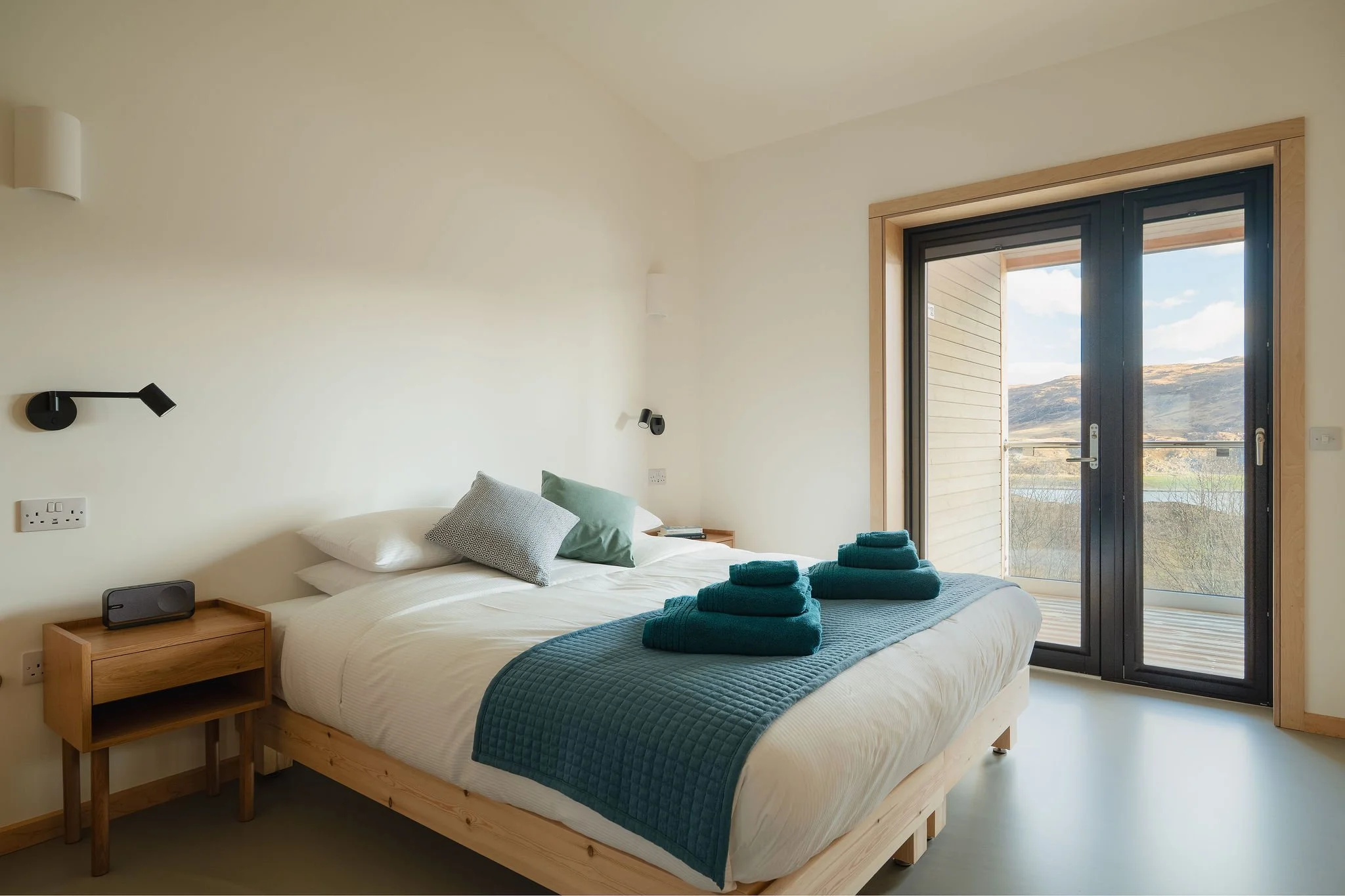 Ein modernes Schlafzimmer mit einem Bettgestell aus Holz, weißer Bettwäsche und blaugrünen Decken. Auf dem Bett liegen zwei aufgerollte blaugrüne Handtücher. Der Raum verfügt über große Glasschiebetüren, die auf einen Balkon mit Blick auf eine hügelige Landschaft führen. Ein Nachttisch mit einem Lautsprecher 