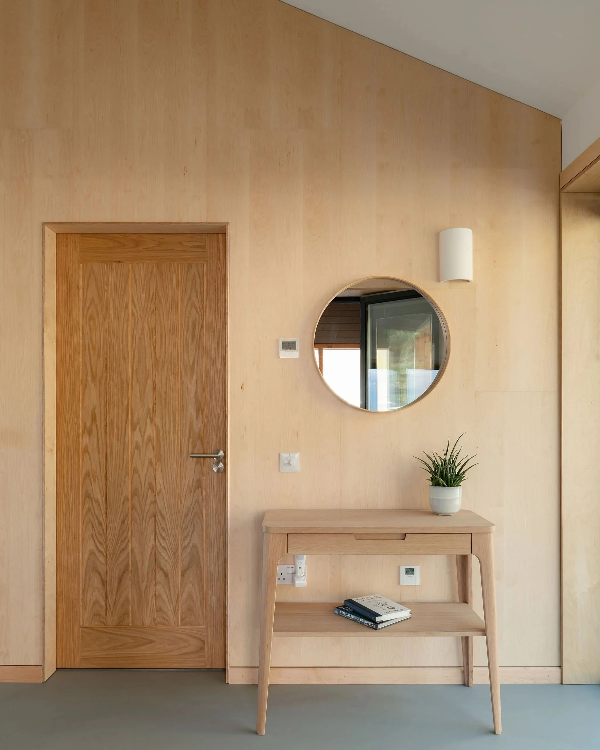 Interior scene featuring a wooden door, a round mirror, a white wall sconce, and a light wood table with a potted plant and books.