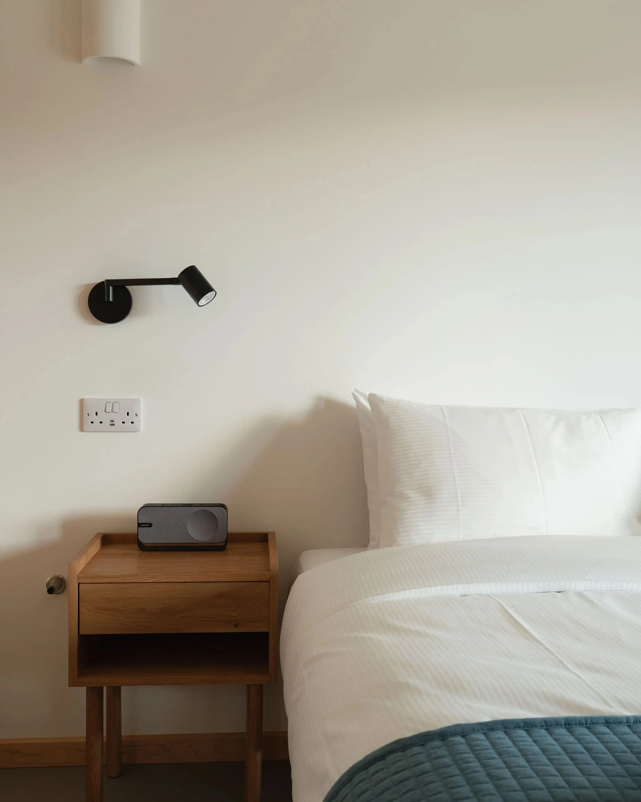 A bedroom with a wooden nightstand next to a bed with white bedding, there is a black speaker on the nightstand, a white wall with a power outlet, and a black wall-mounted reading lamp.
