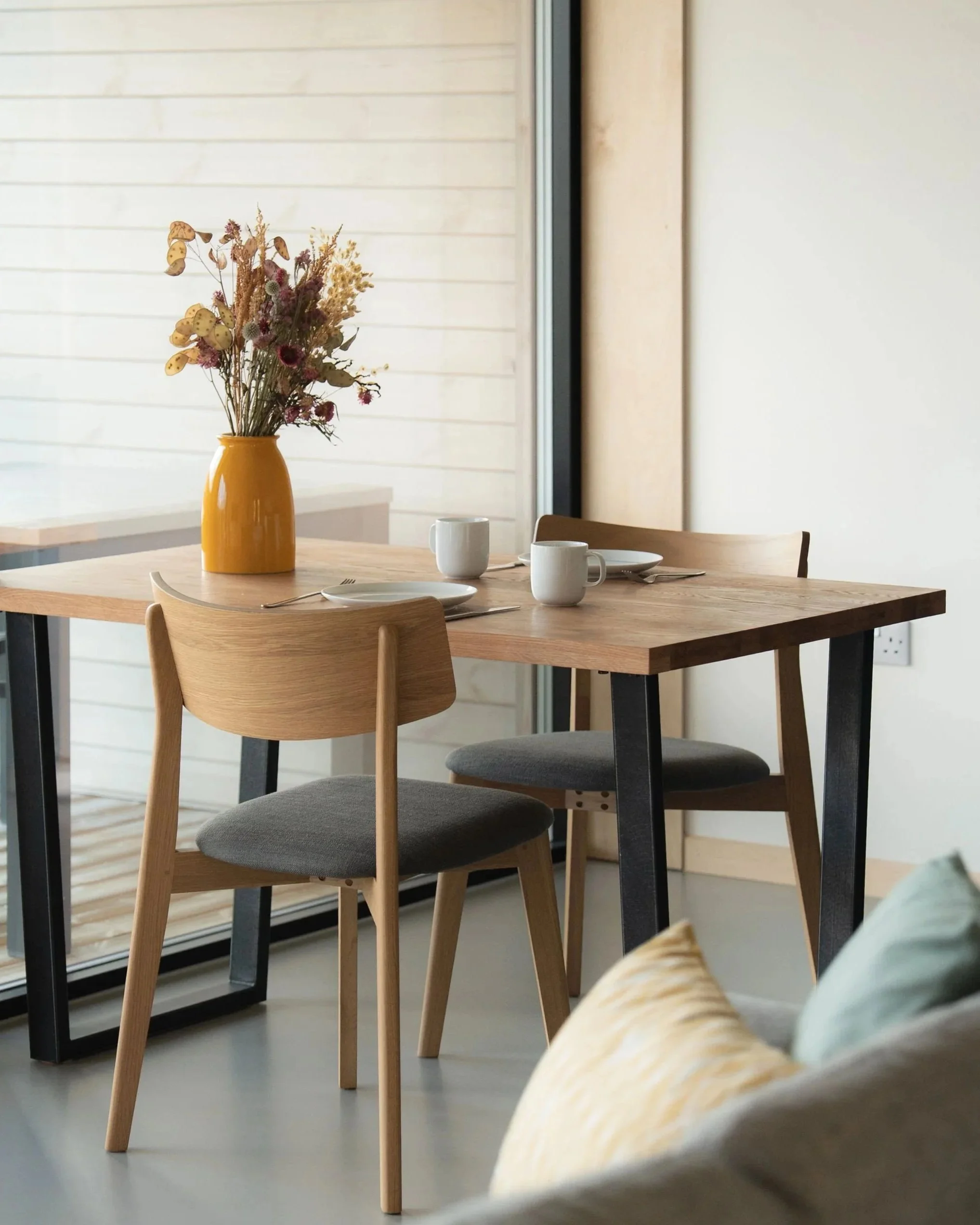 A dining table set with three white cups on white plates and a yellow vase with dried flowers, with wooden chairs around the table in a bright room.