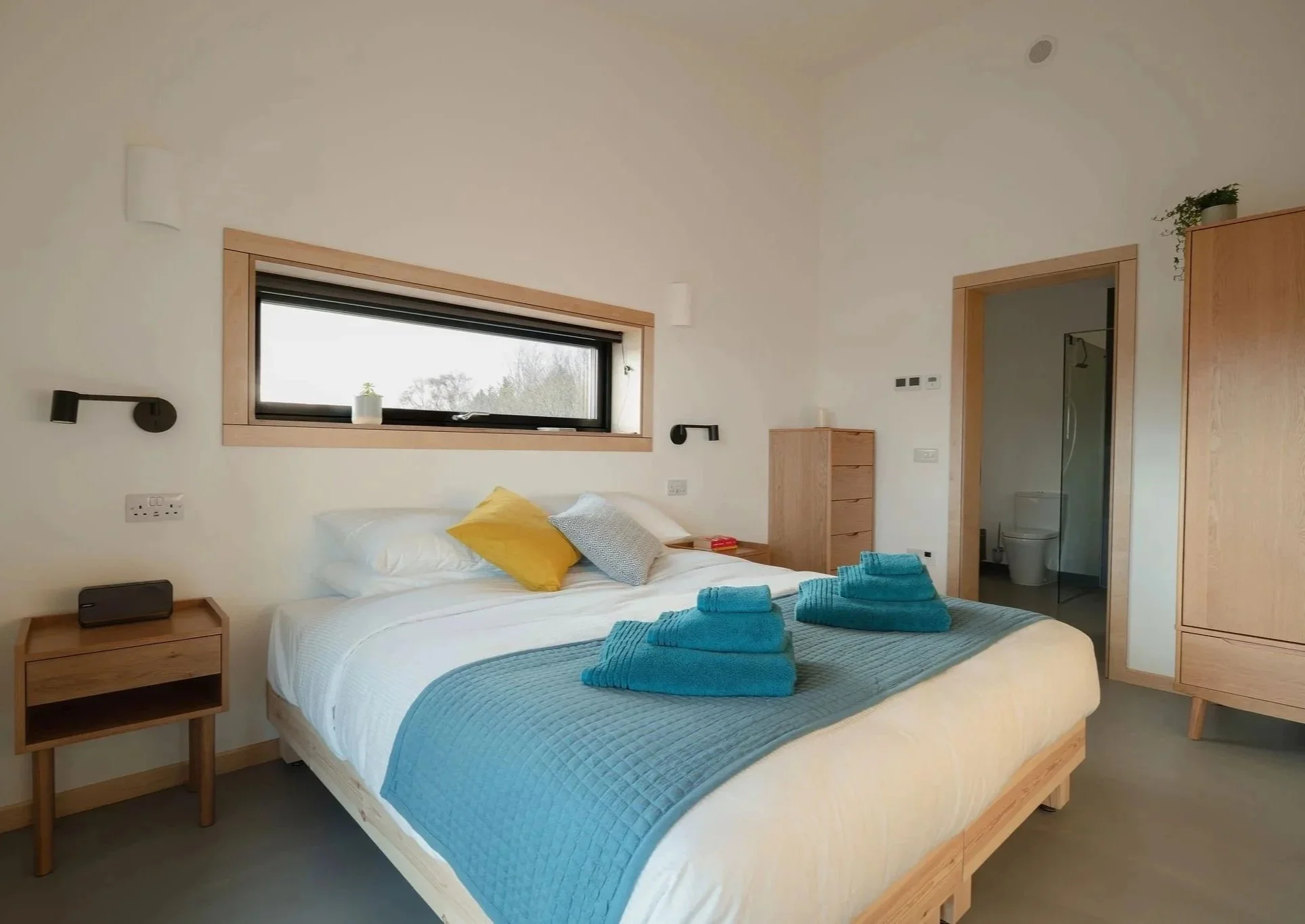 Ein modernes Schlafzimmer mit weißen Wänden, Holzmöbeln, einem Bett mit blauer Bettwäsche und gelben sowie grauen Kissen, türkisfarbenen Handtüchern am Fußende des Bettes, einem Fenster mit einer kleinen Pflanze und einem angrenzenden Badezimmer.