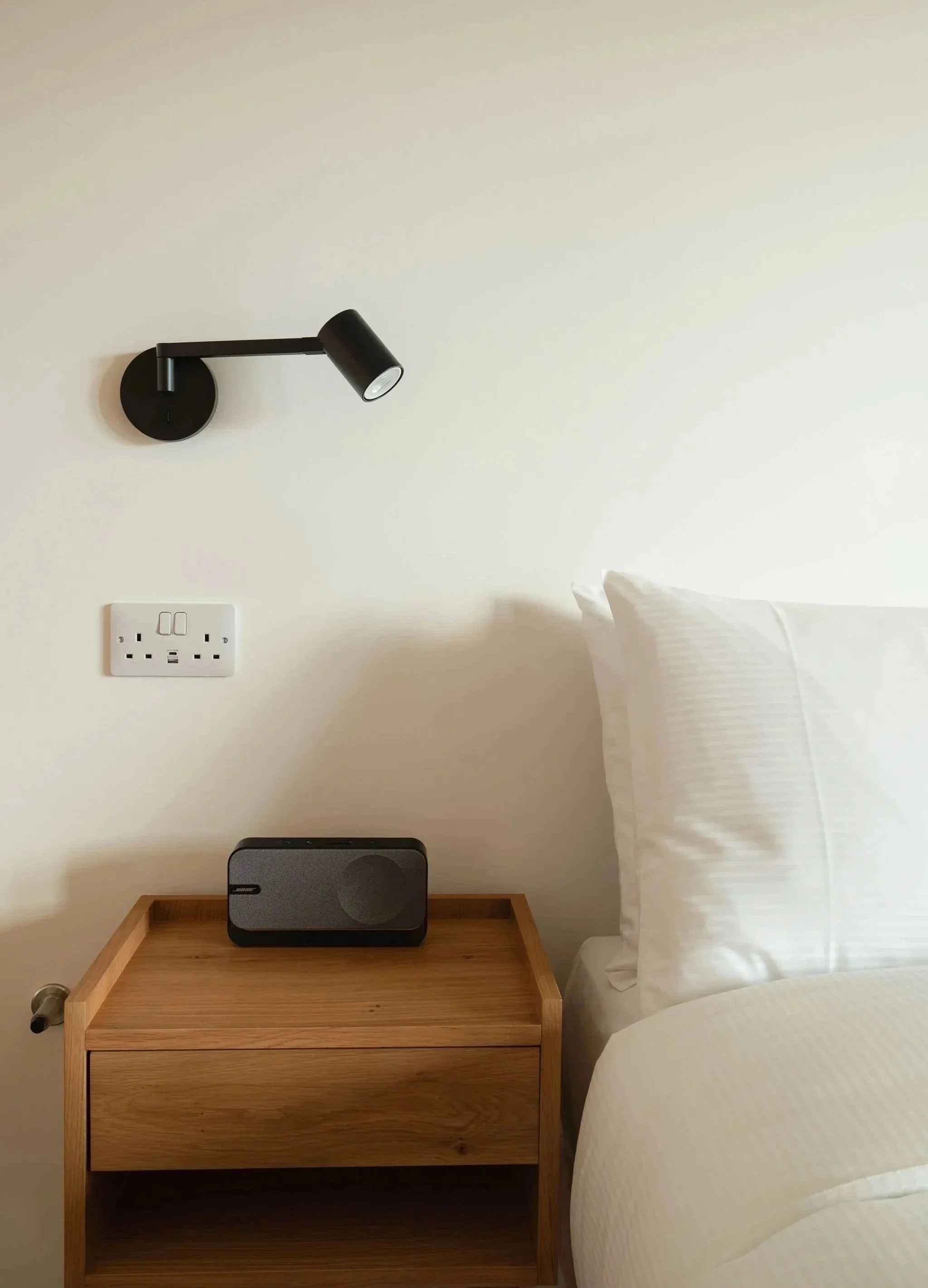 Ein Schlafzimmer mit einer weißen Wand, einer schwarzen, verstellbaren Wandleuchte, einer Steckdose, einem Nachttisch aus Holz, einem Bose-Lautsprecher, einem Bett mit weißer Bettwäsche und einem Kissen.