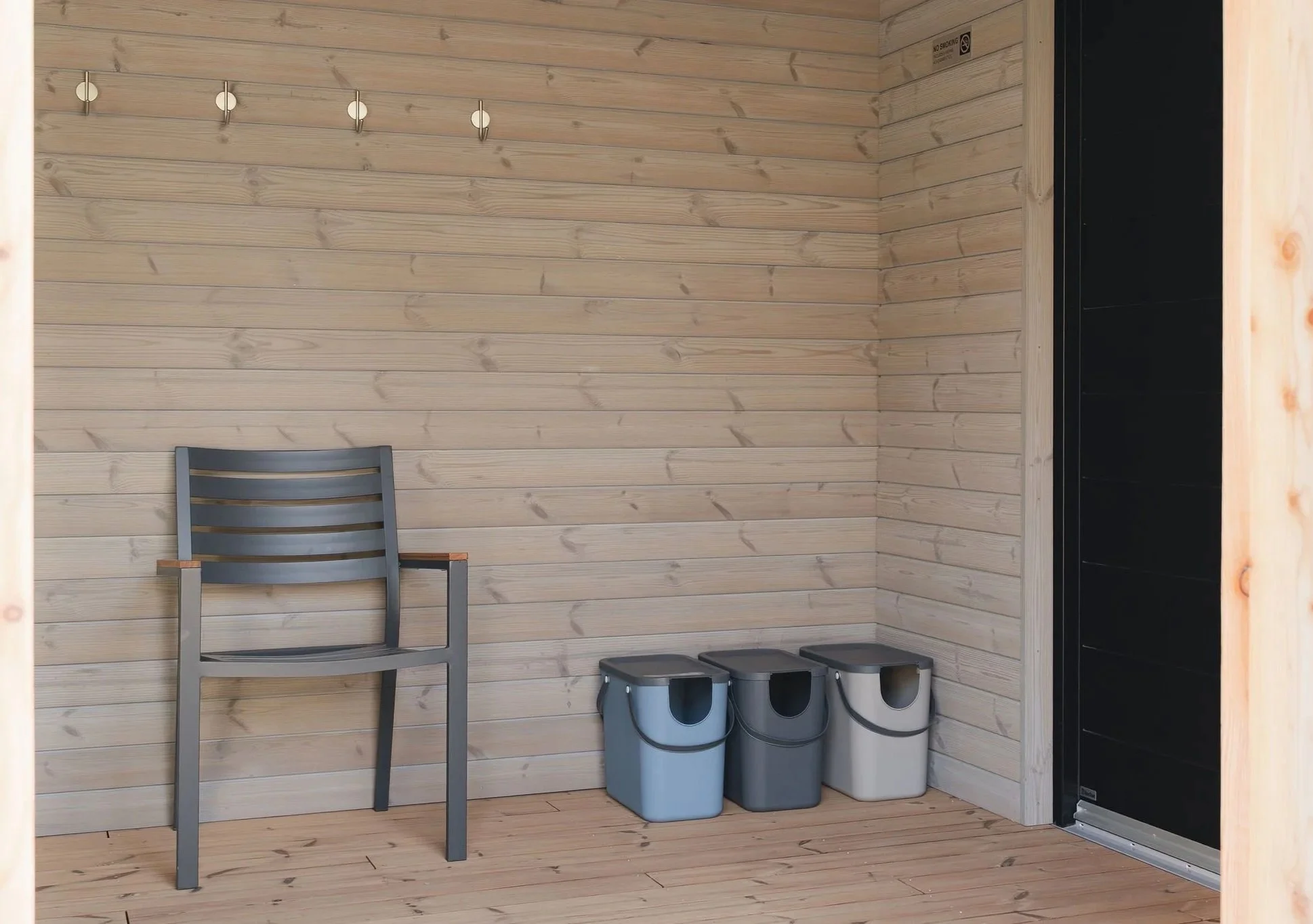 Eine Ecke eines Raums mit hellen Holzwänden und -böden. Dort stehen ein grauer Stuhl, drei kleine graue Hocker und zwei graue Mülleimer. Rechts ist ein Teil einer schwarzen Tür oder Wand zu sehen. An der Wand ist ein Kleiderhaken oder eine Garderobe mit mehreren Haken angebracht, und ein
