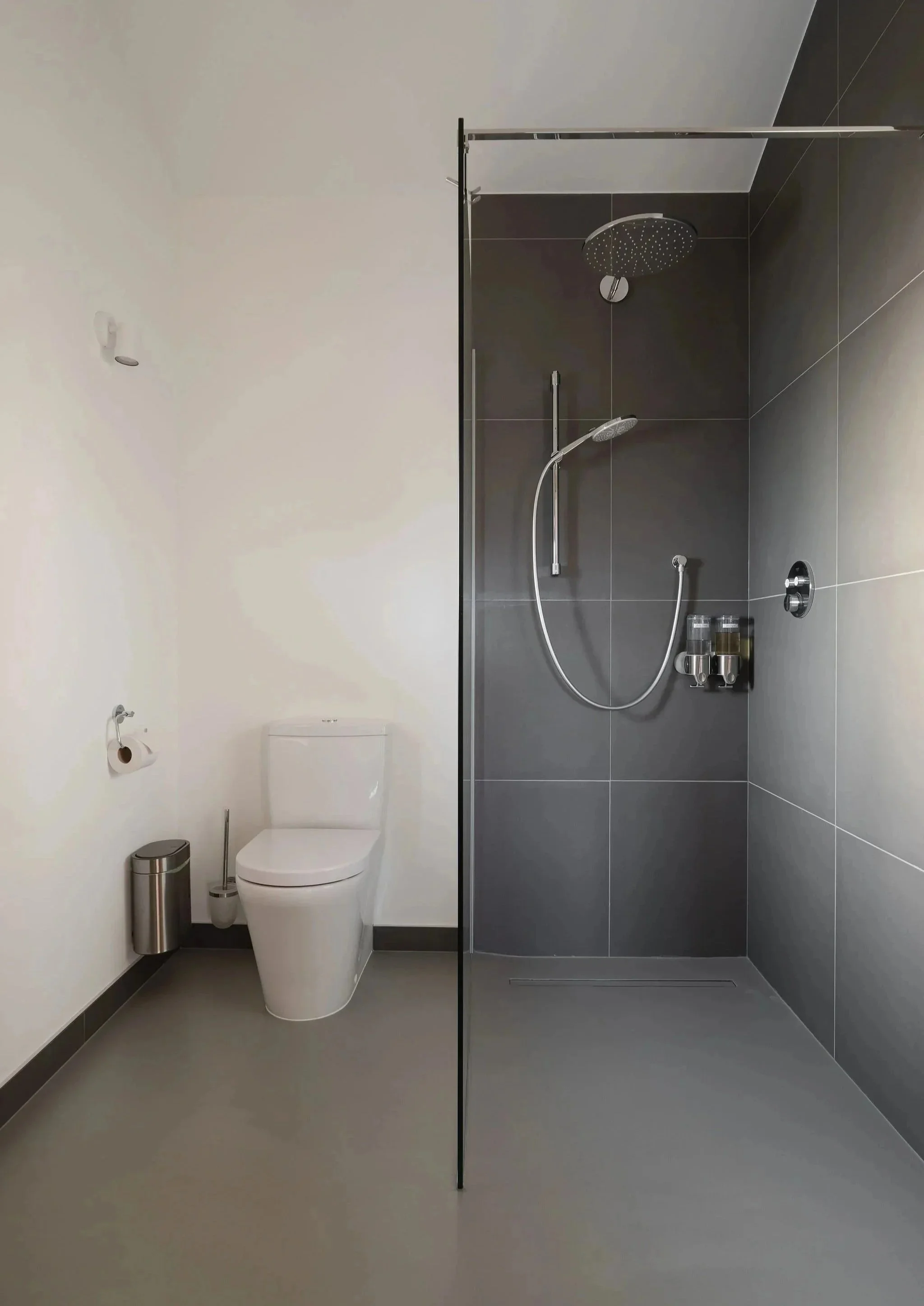Minimalistisches Badezimmer mit einer weißen Toilette, einem wandmontierten Toilettenpapierhalter, einem kleinen Mülleimer, einem Duschbereich mit Glaswand, einem großen runden Regenduschkopf, einer Handbrause und einer dunkel gefliesten Wand, auf der einige Flaschen mit Pflegeprodukten stehen 