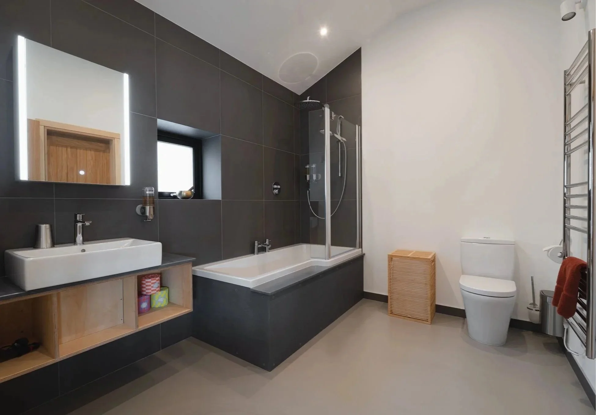 Modernes Badezimmer mit schwarz gefliesten Wänden, weißen Sanitäranlagen, einer Badewanne mit verstellbarer Dusche, einem Bidet mit Waschbecken, einer Toilette, einem Spiegel mit LED-Beleuchtung, einem kleinen Fenster, einem Wäschekorb aus Korbgeflecht und Badezimmeraccessoires.