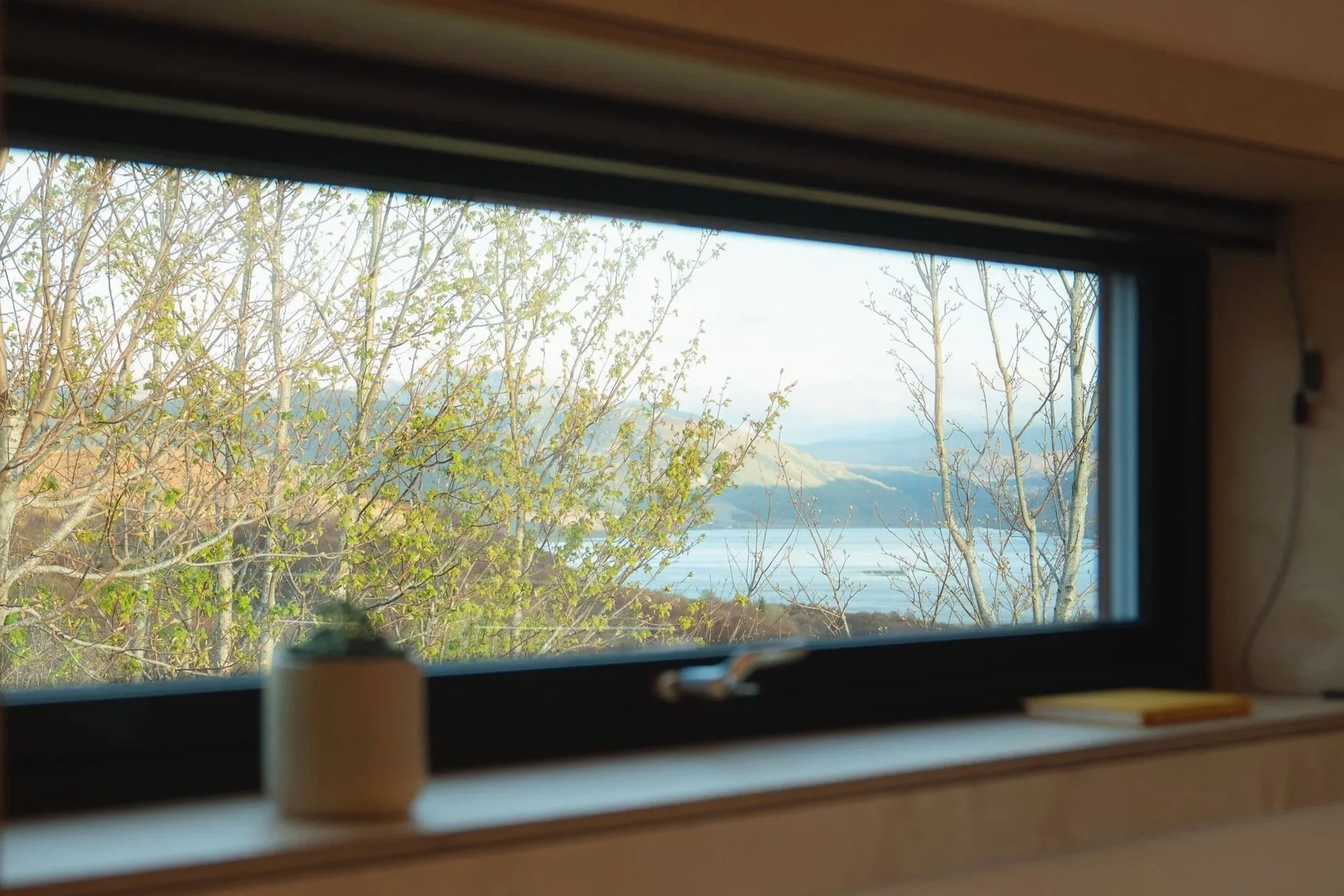 Blick durch ein Fenster auf Bäume mit knospenden Blättern, ein Gewässer und entfernte Hügel oder Berge, eingerahmt von einem hölzernen Fensterrahmen.