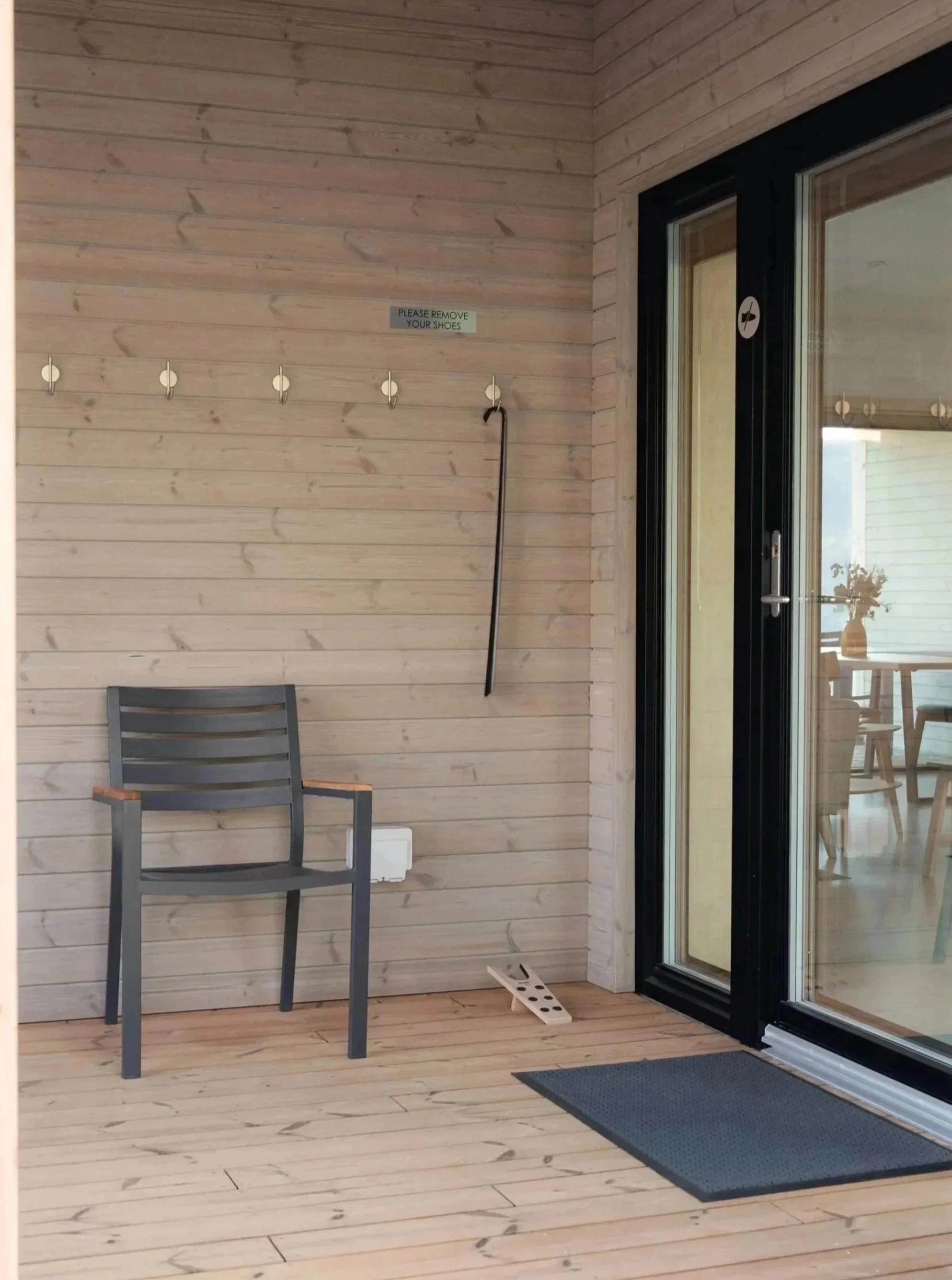 Eine Holzveranda mit einem Stuhl, einem Schild mit der Aufschrift „Bitte Schuhe ausziehen“, einem an der Wand hängenden Schuhlöffel, einer schwarzen Tür mit einem Aufkleber, der darauf hinweist, dass Schuhe nicht erlaubt sind, einer Fußmatte und einer Glasschiebetür, die ins Haus führt.