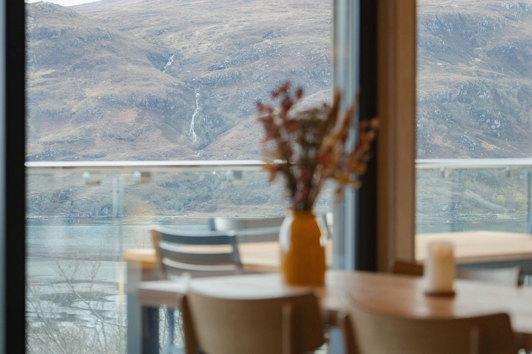 View of a mountain landscape through a window, with a blurred table and chairs inside the room, and a vase with dried flowers on the windowsill.