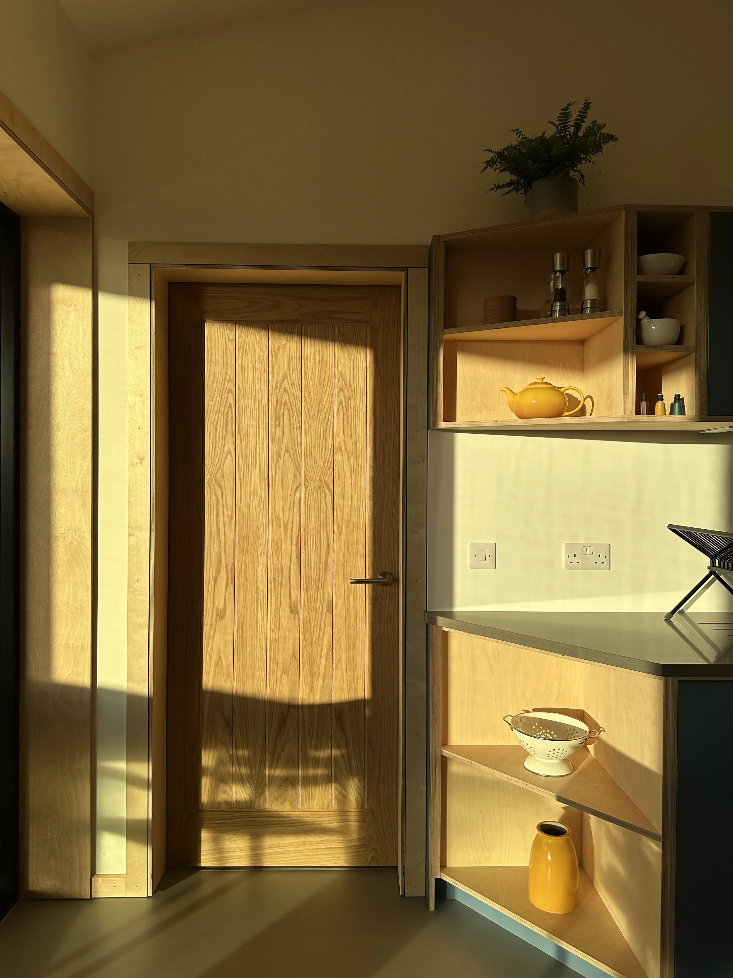 A wooden kitchen cabinet with open shelves displaying white bowls, a yellow teapot, spice jars, and a decorative plant on top. There is electrical outlet on the backsplash and sunlight casting shadows on the door and wall.