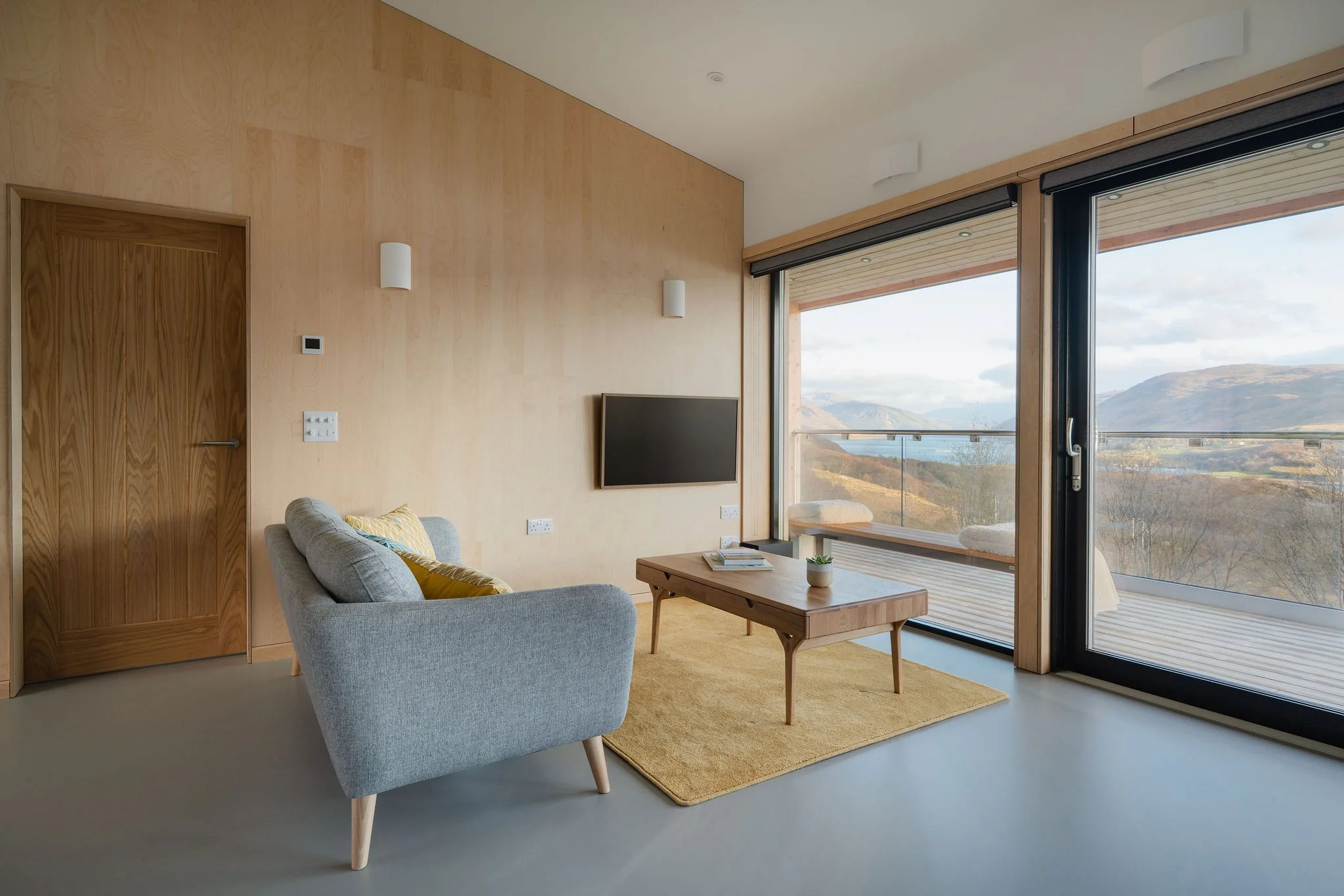 Ein modernes Wohnzimmer mit einem grauen Sessel, einem Couchtisch aus Holz, einem gelben Teppich, einem an der Wand montierten Fernseher, großen Fenstern mit Blick auf eine Berglandschaft und einer Holztür auf der linken Seite.
