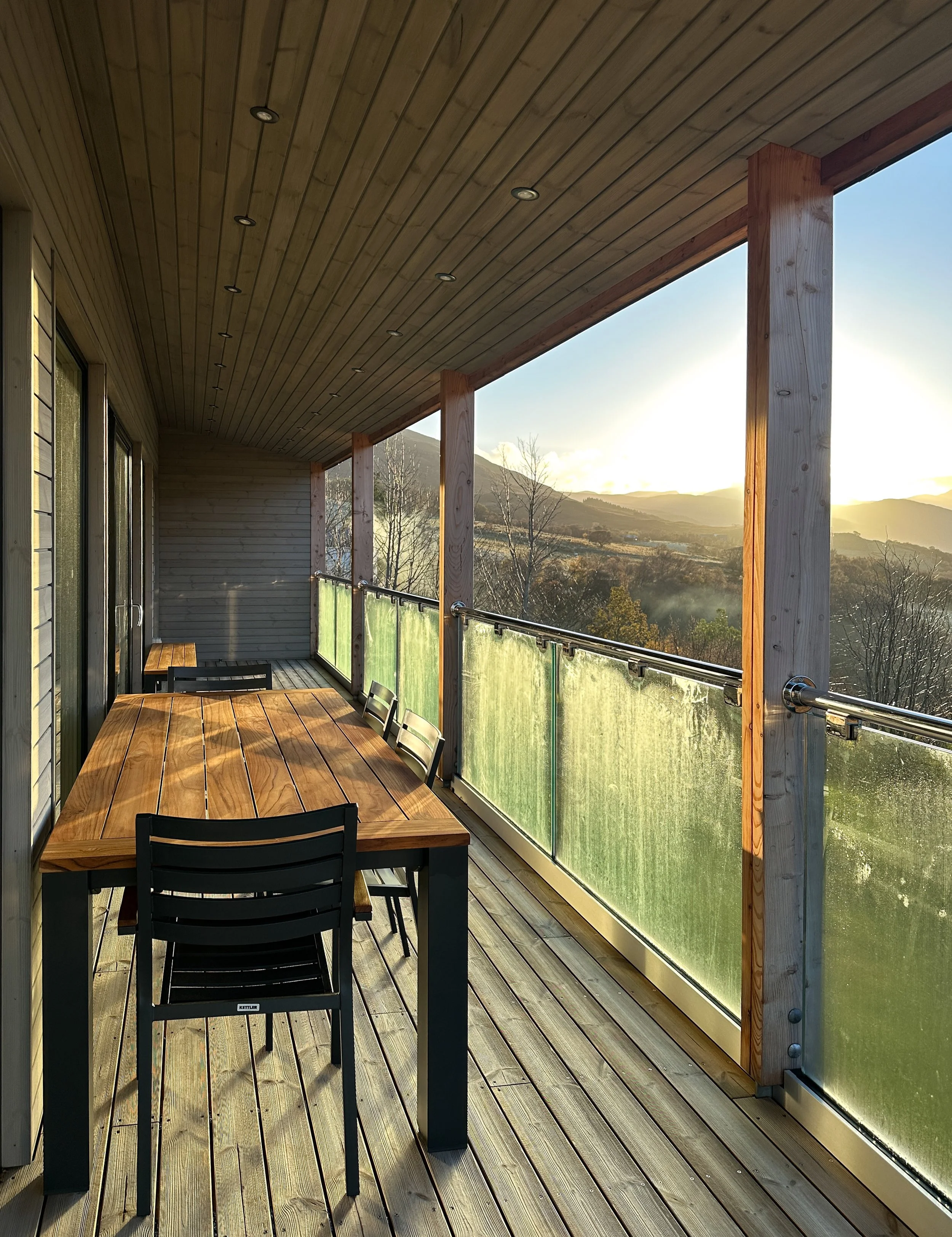 Balcony with wooden floor, table, and chairs overlooking a scenic landscape with trees and mountains during sunset.
