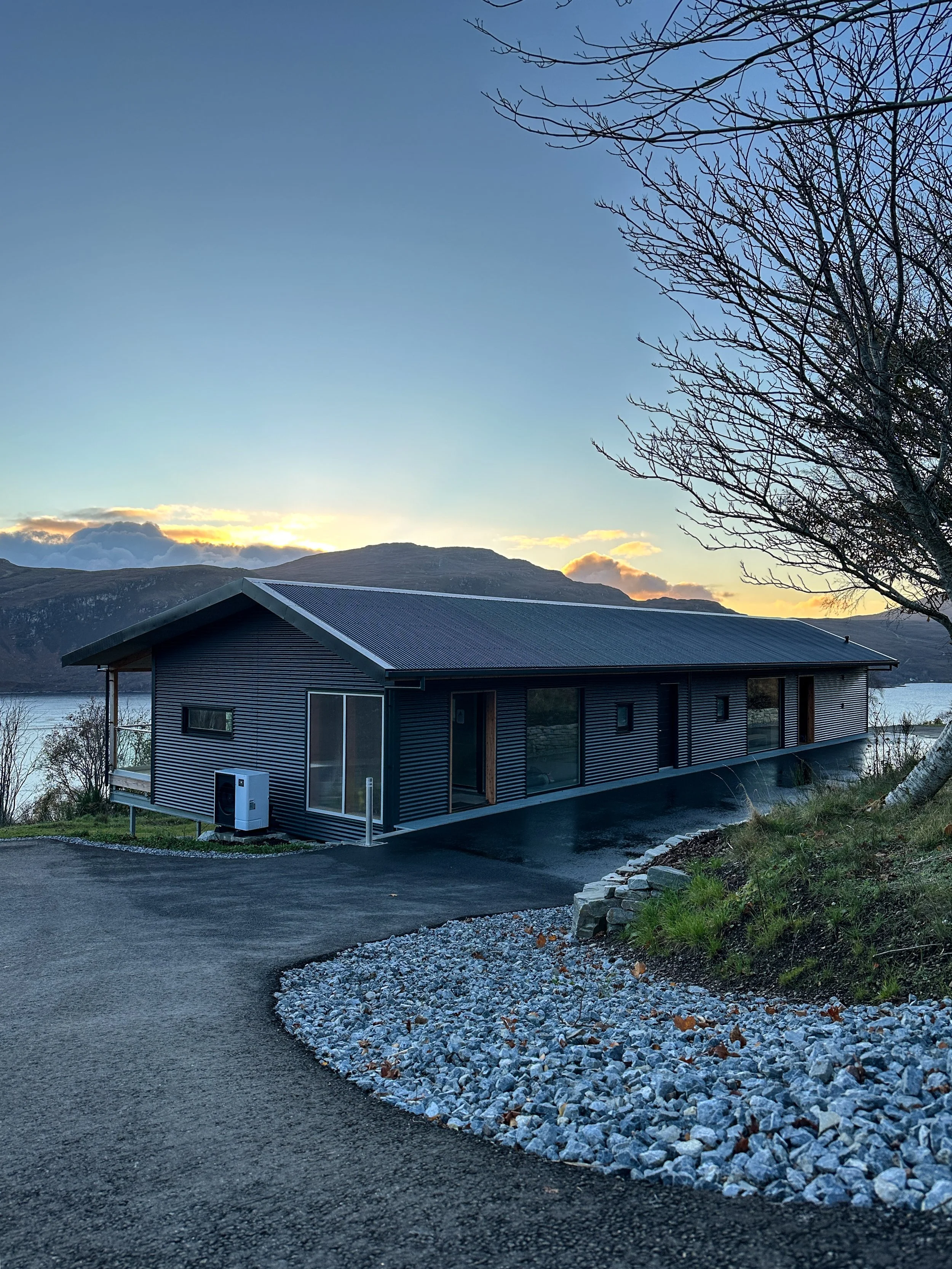 Modern single-story house with dark siding, large windows, and a sloped metal roof, situated near a body of water with mountains in the background at sunset.
