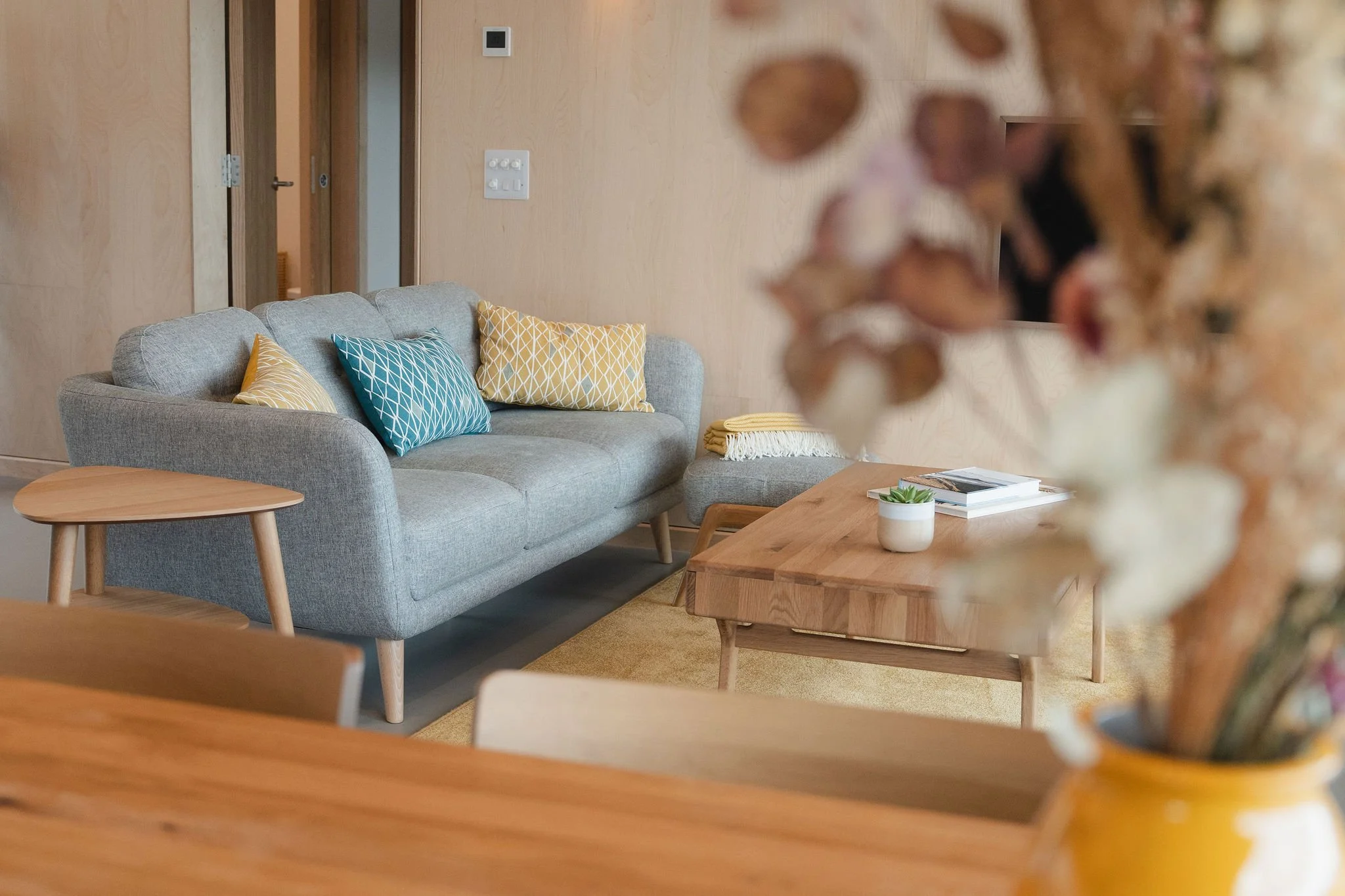 Wohnzimmer mit einem grauen Sofa, auf dem gelbe und blaue gemusterte Kissen liegen, einem Couchtisch aus Holz mit einer kleinen Pflanze und Zeitschriften sowie einem unscharfen Blumenstrauß im Vordergrund.