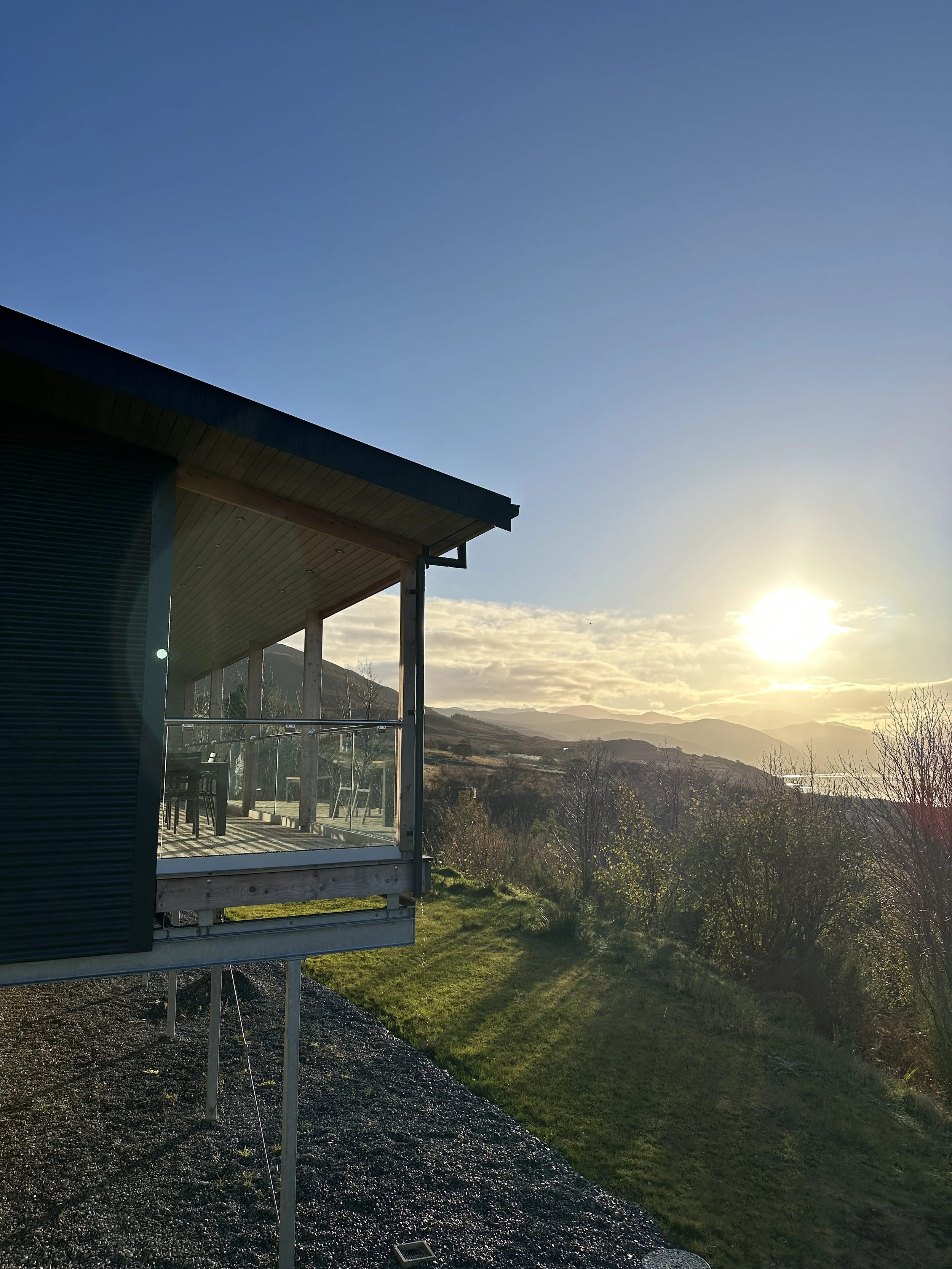 A modern house with a balcony overlooking a grassy hillside and distant mountains during sunset.