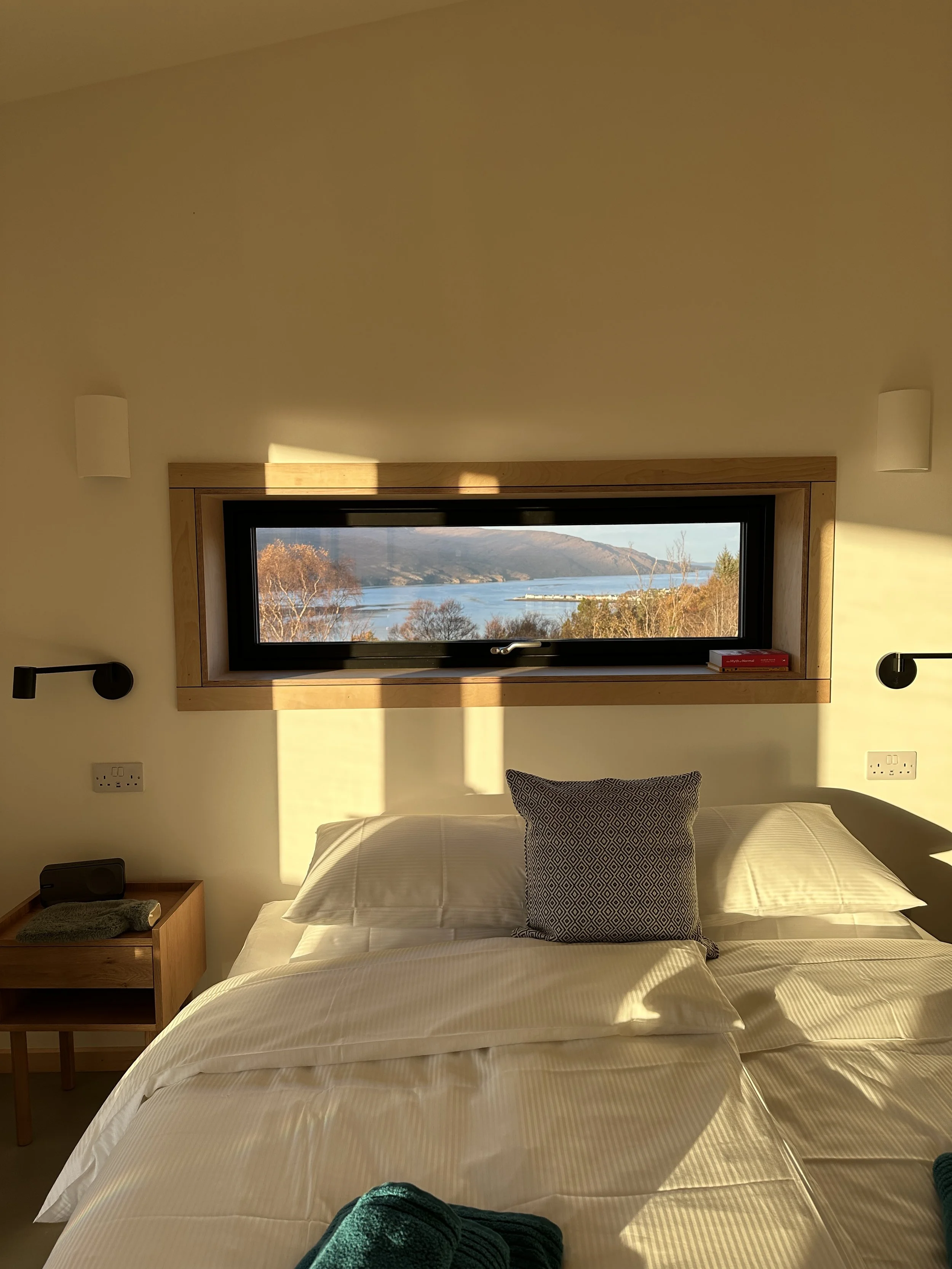 A cozy bedroom with a large bed, white bedding, and a decorative pillow. A small wooden nightstand with a speaker and a book. A horizontal window above the bed offers a scenic view of the sea, trees, and mountains during sunset.