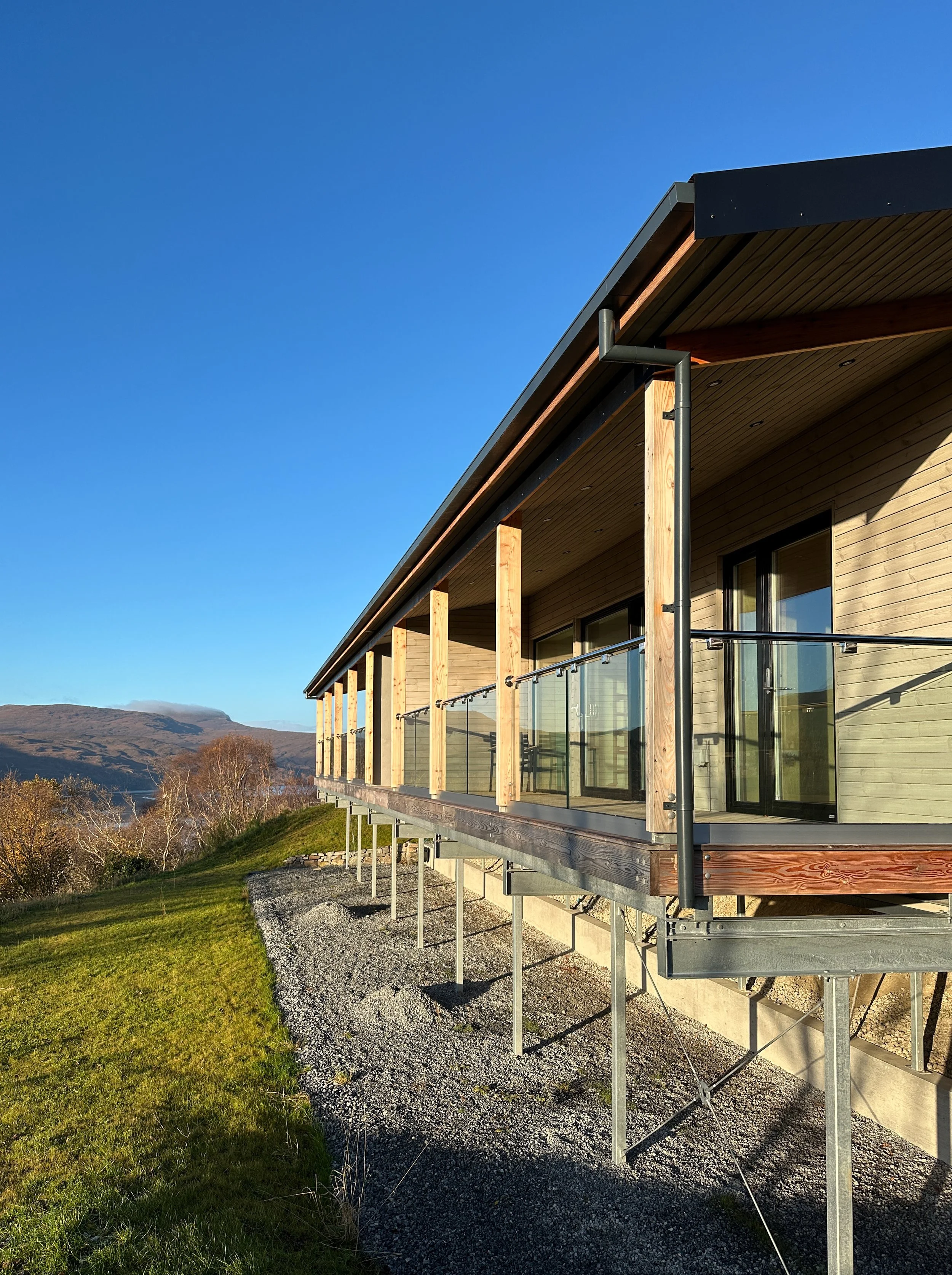 A modern house with a wooden exterior and glass balcony railing, elevated on metal supports, set against a hillside landscape under a clear blue sky.