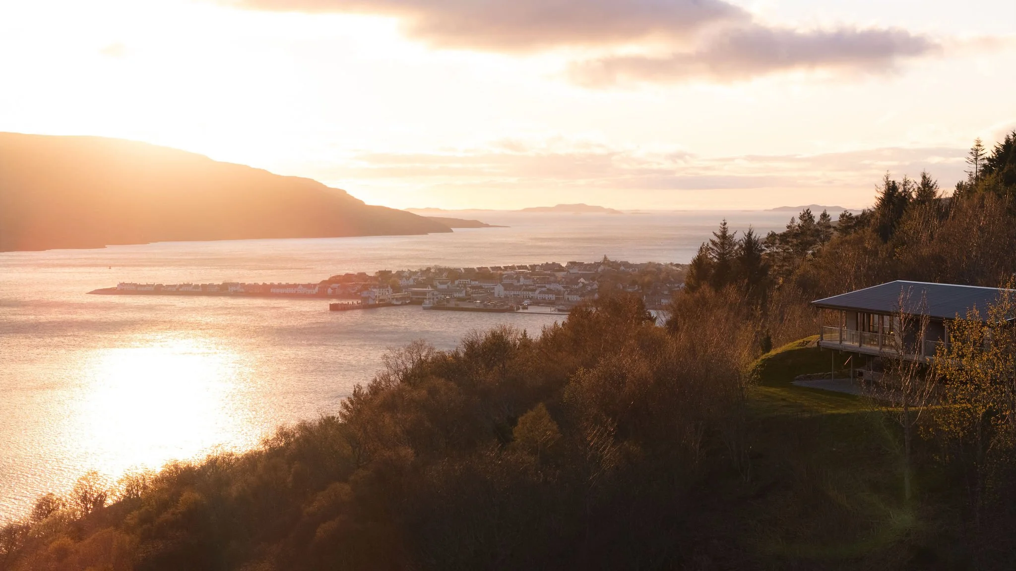 Sunset over a coastal town with buildings along the shoreline, hills, and trees, and a modern house on a hill in the foreground.
