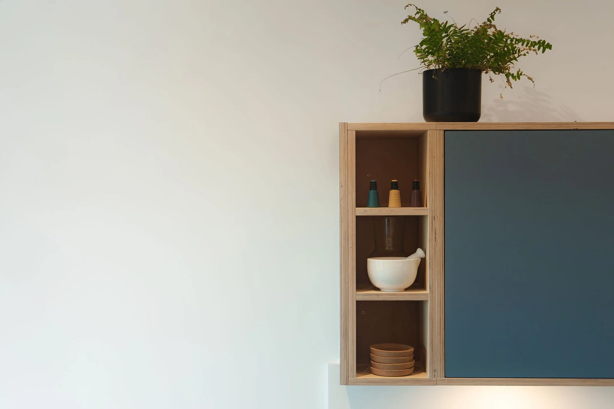 A wooden shelving unit with open compartments, a white bowl, and stacked brown bowls, set against a background with half white and half dark blue wall, with a potted plant on top.