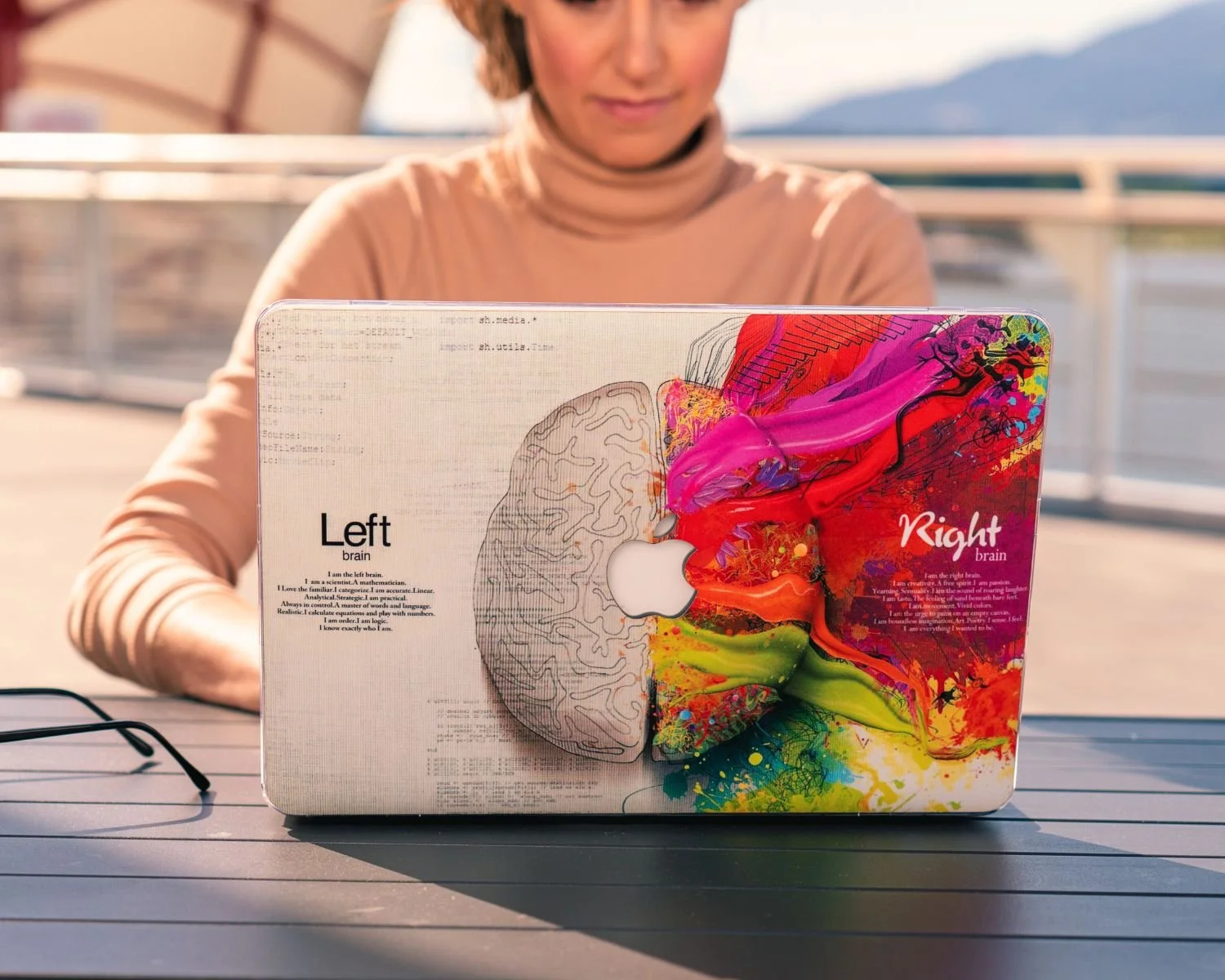 A person sitting outdoors at a table with a colorful sticker on a laptop, featuring a split image of a brain diagram labeled 'Left' and 'Right' with vibrant paint splashes.