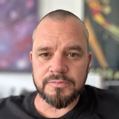Close-up of a man with a shaved head and beard, looking directly at the camera inside a room.