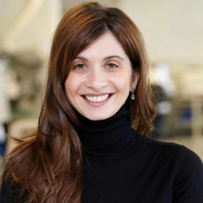A woman with long, wavy auburn hair, wearing a black turtleneck sweater, smiling in an indoor setting.