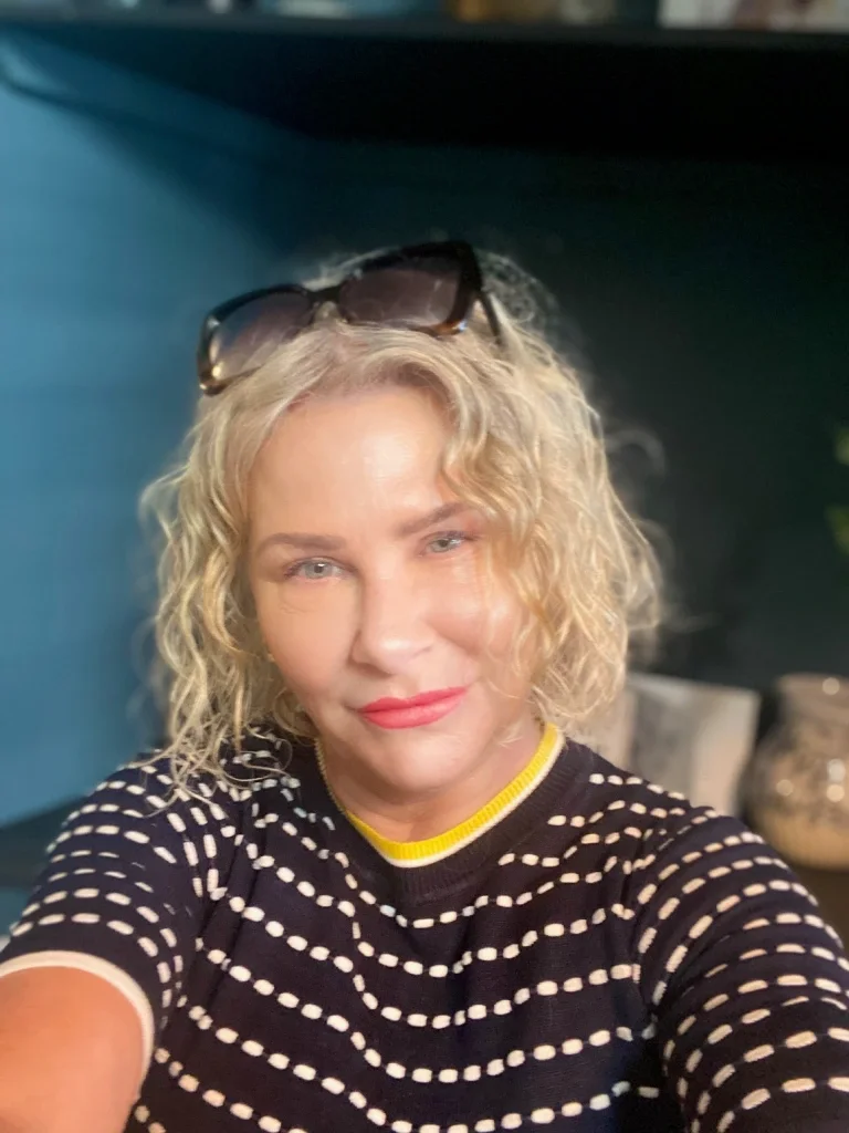 A woman with blonde curly hair, wearing sunglasses on her head and a black and white striped shirt, taking a selfie.