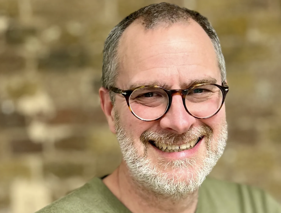 A smiling middle-aged man with short gray hair, glasses, and a beard, wearing a green shirt, aged indoors with a blurred brick wall background.