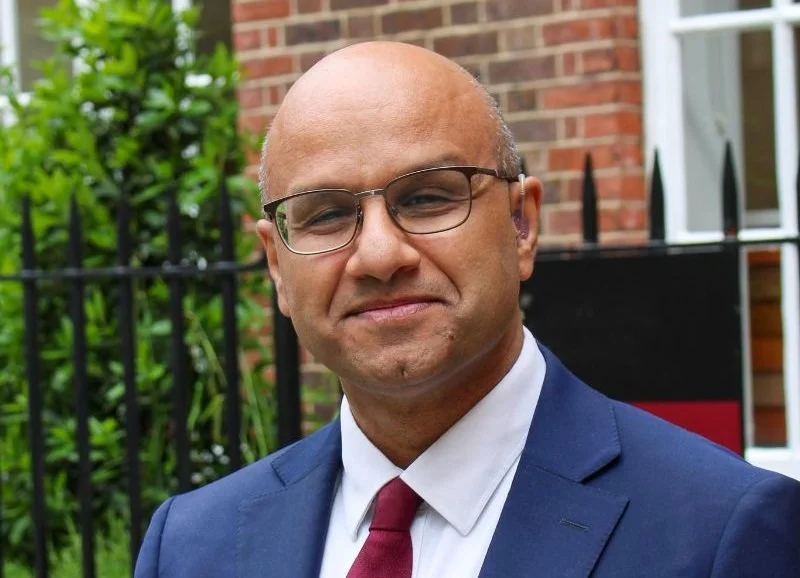 A man in a blue suit, white shirt, and red tie standing outdoors in front of a brick building and greenery.